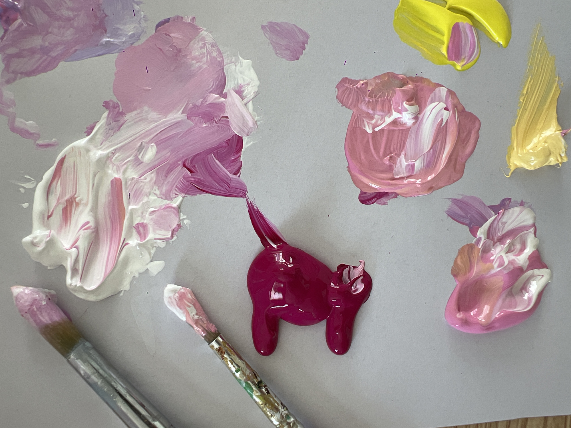 A flat lay of paint colors on a light gray surface shows two paint brushes and several dollops of acrylic paint. The brushes are positioned in the upper left quadrant. The larger brush, angled down and to the right, has pink and white paint on its bristles. Below it, a smaller brush, also angled down and to the right, displays pink and white paint.
The paint dollops are distributed across the surface. In the upper portion, a large swirl of white and pink paint dominates, with hints of light purple in the upper right corner. Below this, a bright, concentrated dollop of magenta paint is prominent in the middle left. Further down and to the right, a textured dollop of pink and white paint is visible. To its right, a bright yellow dollop with streaks of pink and white is present. Finally, in the lower left, another swirl of pink, white, and light purple paint is depicted, and in the bottom right, a streak of yellow paint is visible. The overall impression is one of an artist's palette with various color combinations.
The image does not contain any elements that would indicate a specific location in the city of Bac Nha Trang Ward, Vietnam.