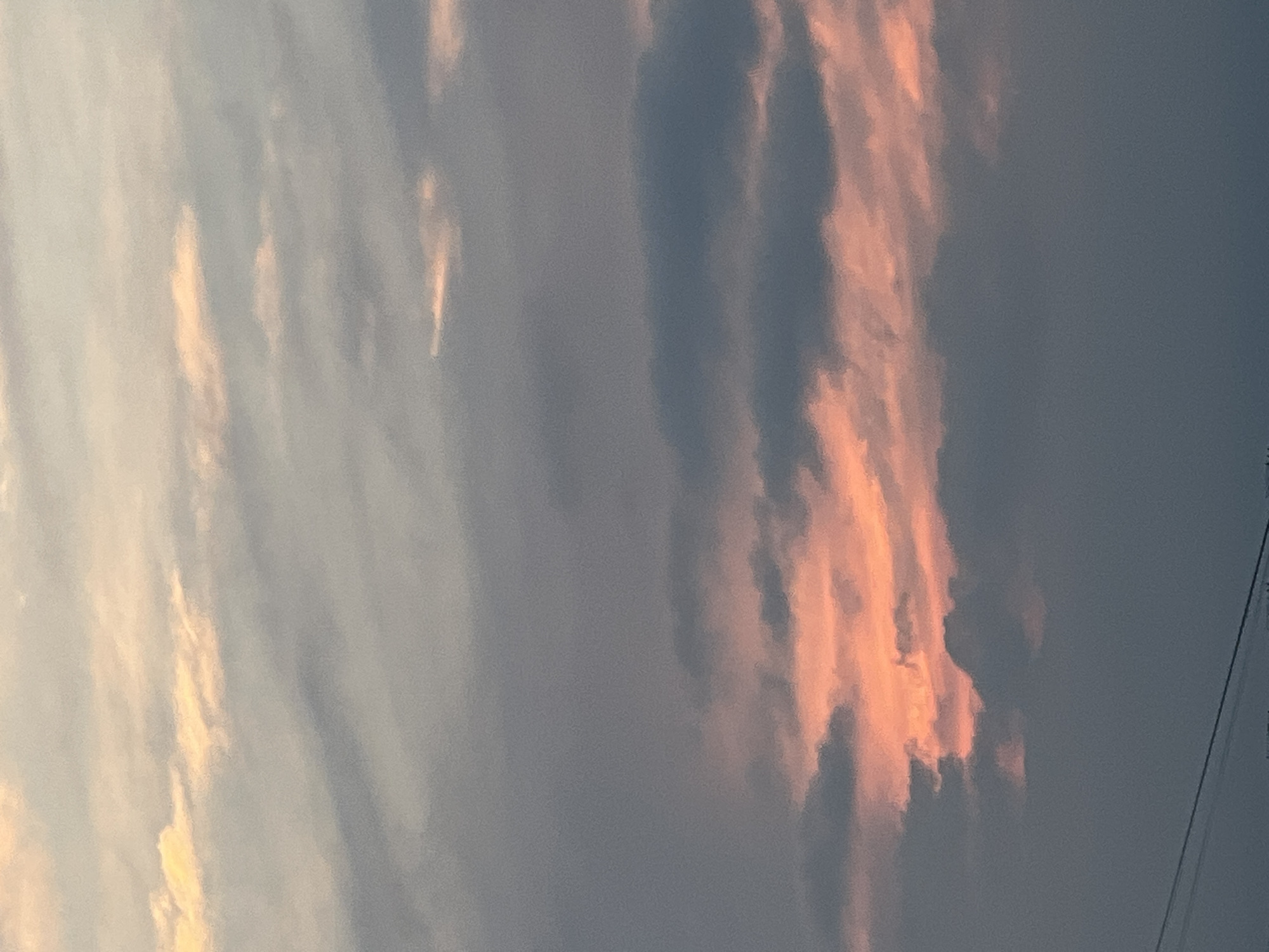 An outdoor, eye-level, medium shot captures a dusk sky filled with clouds. The clouds are a mix of gray and pink, with the pink hues suggesting the last rays of sunlight. The sky is vast and covers the entire frame, with a thin line of telephone wires visible at the bottom of the image. No people or objects are discernable.