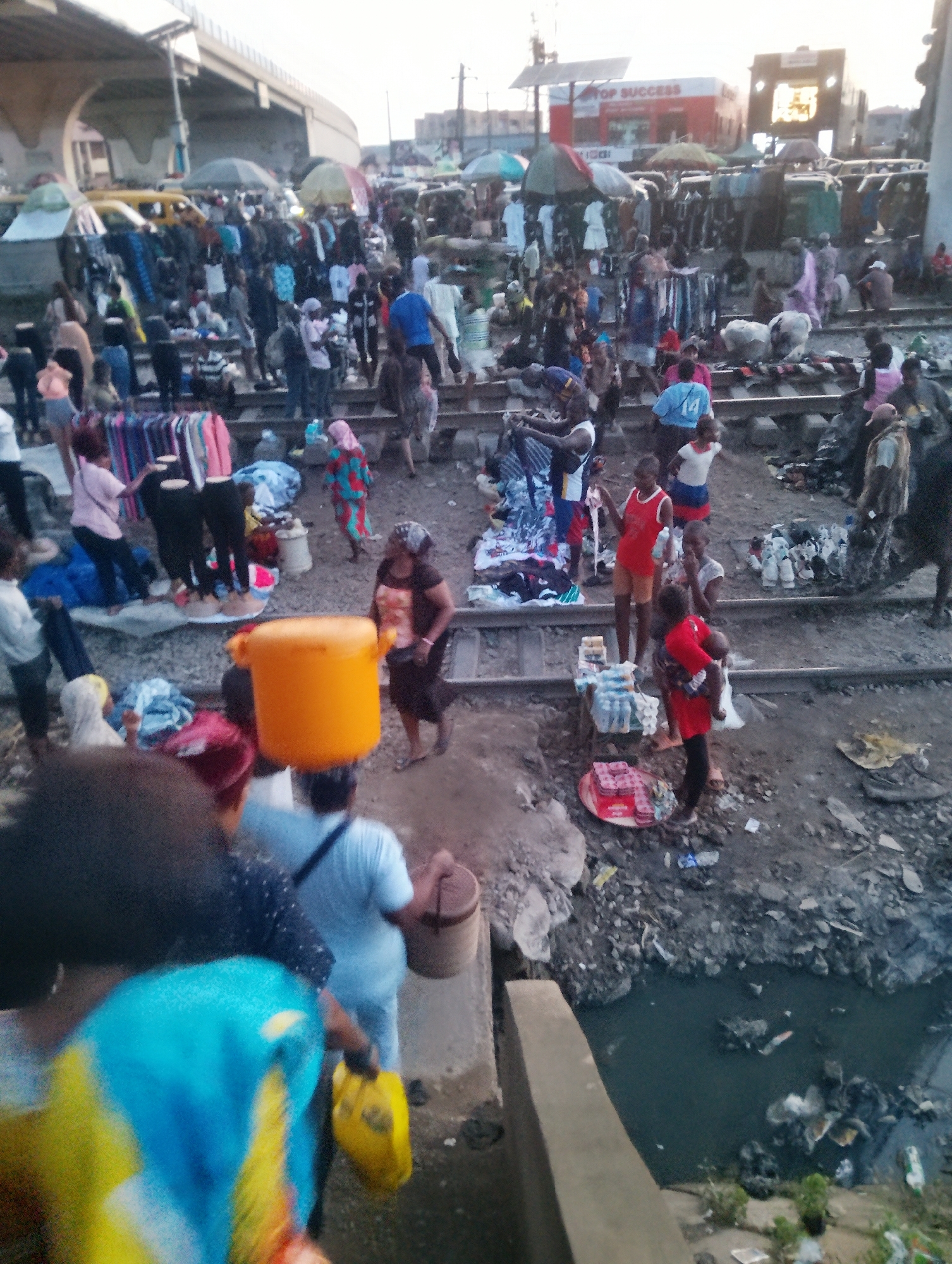 This image depicts a busy open-air market set up along railway tracks, likely in Ojokoro, Nigeria. Numerous people are engaged in various activities, suggesting a vibrant and active commercial scene. The market appears crowded with individuals browsing goods, interacting with vendors, and moving through the pathways.
The subjects are diverse, including men, women, and children, all dressed in everyday clothing. Objects prominent in the scene are stalls filled with clothing, textiles, and other merchandise. Several individuals are carrying items, such as a woman with a large orange bucket balanced on her head and another person holding a yellow bag. The ground is littered with debris, and a muddy, dark body of water is visible to the right, possibly a drainage canal. The railway tracks themselves are integrated into the market space, indicating the area's unique layout.
The activity taking place is clearly a market day, characterized by trade and social interaction. People seem to be shopping, selling, and socializing. The overall atmosphere appears lively, though the cluttered environment and the presence of the water body suggest challenging living and working conditions.
Notable details include the time of day, which appears to be late afternoon or early evening, as indicated by the soft, diffused lighting and long shadows. The weather seems to be overcast or hazy. Location cues are the railway tracks and the general look of the marketplace, consistent with many informal markets in Nigeria. There is some visible text on a red and white building in the background, reading "TOP SUCCESS." The image captures a dynamic slice of daily life in a bustling urban or semi-urban environment.