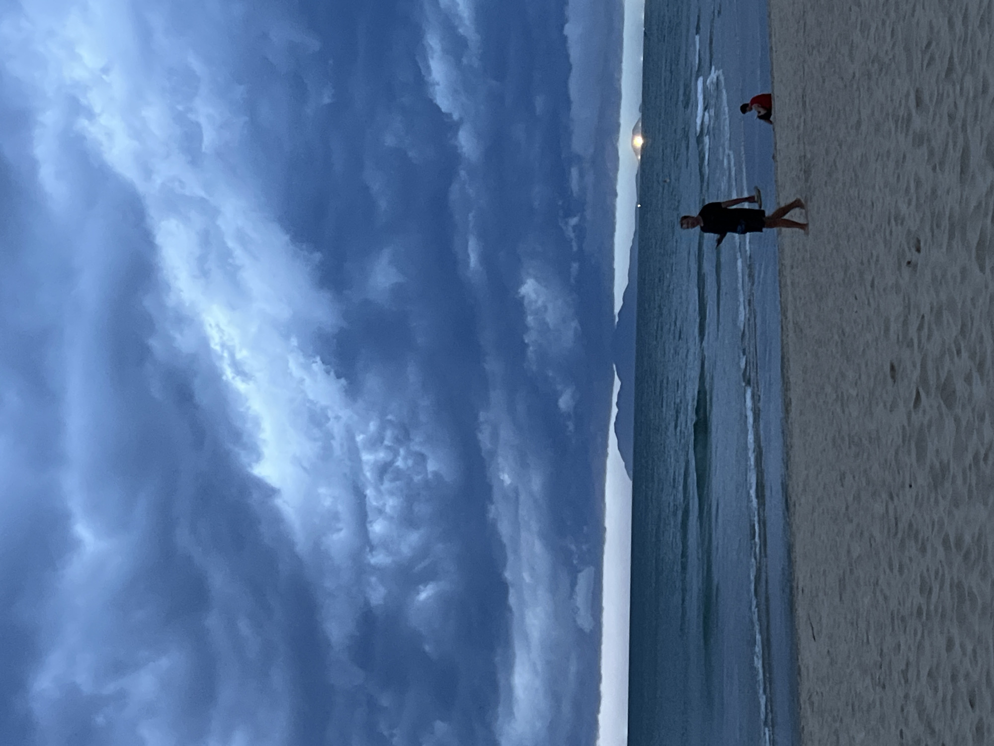 This is a photograph of a beach in Đà Nẵng, Vietnam, taken at dusk. The sky is filled with dark, dramatic clouds, suggesting an impending storm. The sea is calm, with gentle waves lapping at the shore. In the foreground, the sand is light brown and appears somewhat damp.
Two individuals are visible on the beach. Closer to the foreground, a person dressed in a black t-shirt and shorts is walking from left to right. They are carrying a phone in their left hand and a pair of flip-flops in their right. Further back, near the water's edge, another person is seated on the sand, wearing a red top.
In the distance, across the water, there is a hazy silhouette of land, possibly islands or a distant coastline. A bright light, possibly from a boat or a structure, is visible on the horizon. The overall mood of the image is somewhat somber due to the stormy sky, yet also peaceful, with the presence of the individuals on the beach.