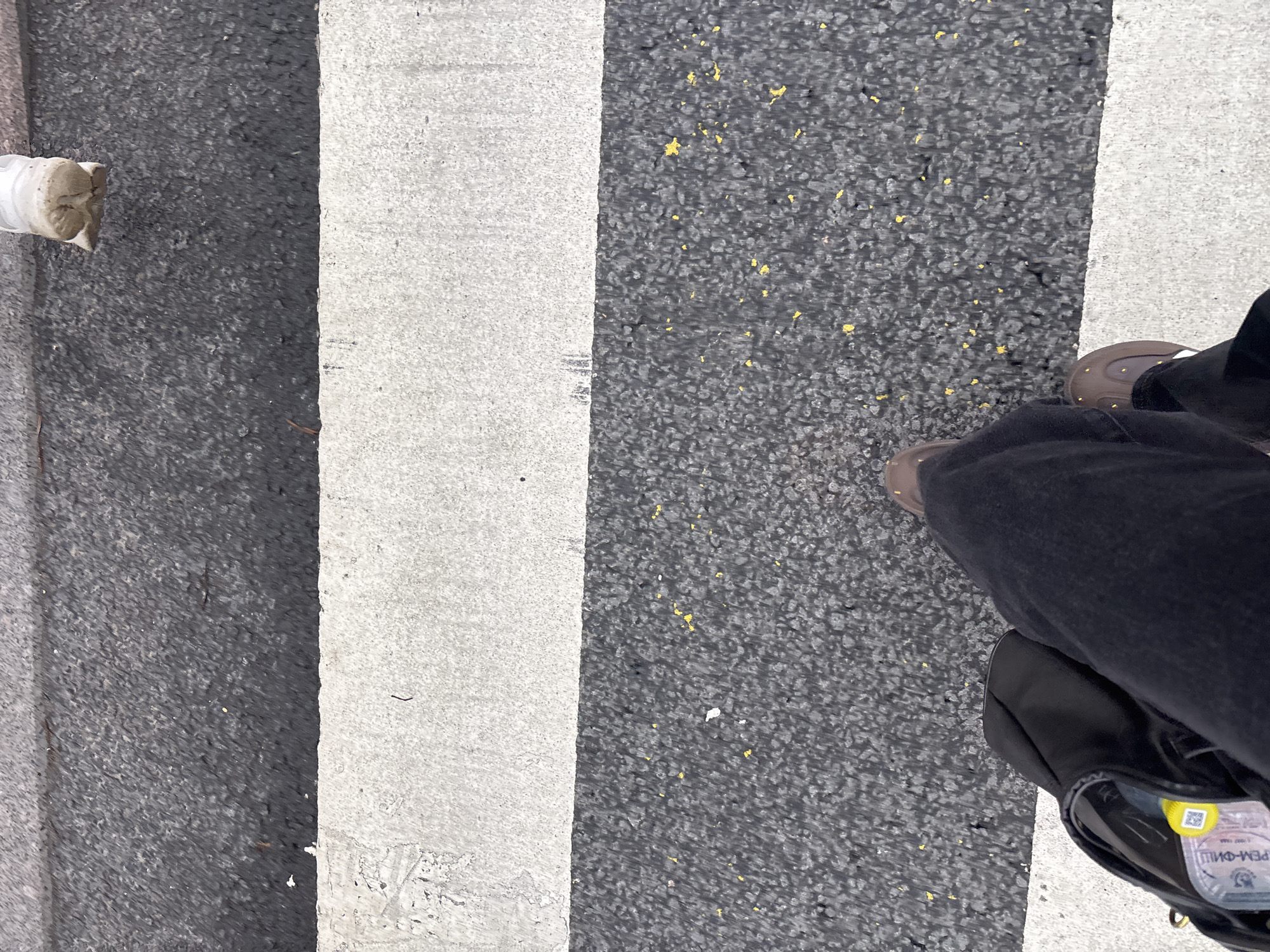 The image shows a person standing on a crosswalk in an urban area. The crosswalk is marked with alternating black asphalt and white painted stripes. The person is wearing brown shoes and black pants, and is carrying a black bag. A discarded paper cup sits at the edge of the frame, left side. Based on the item visible in the bag, the location is identified as Unknown, Russia.
