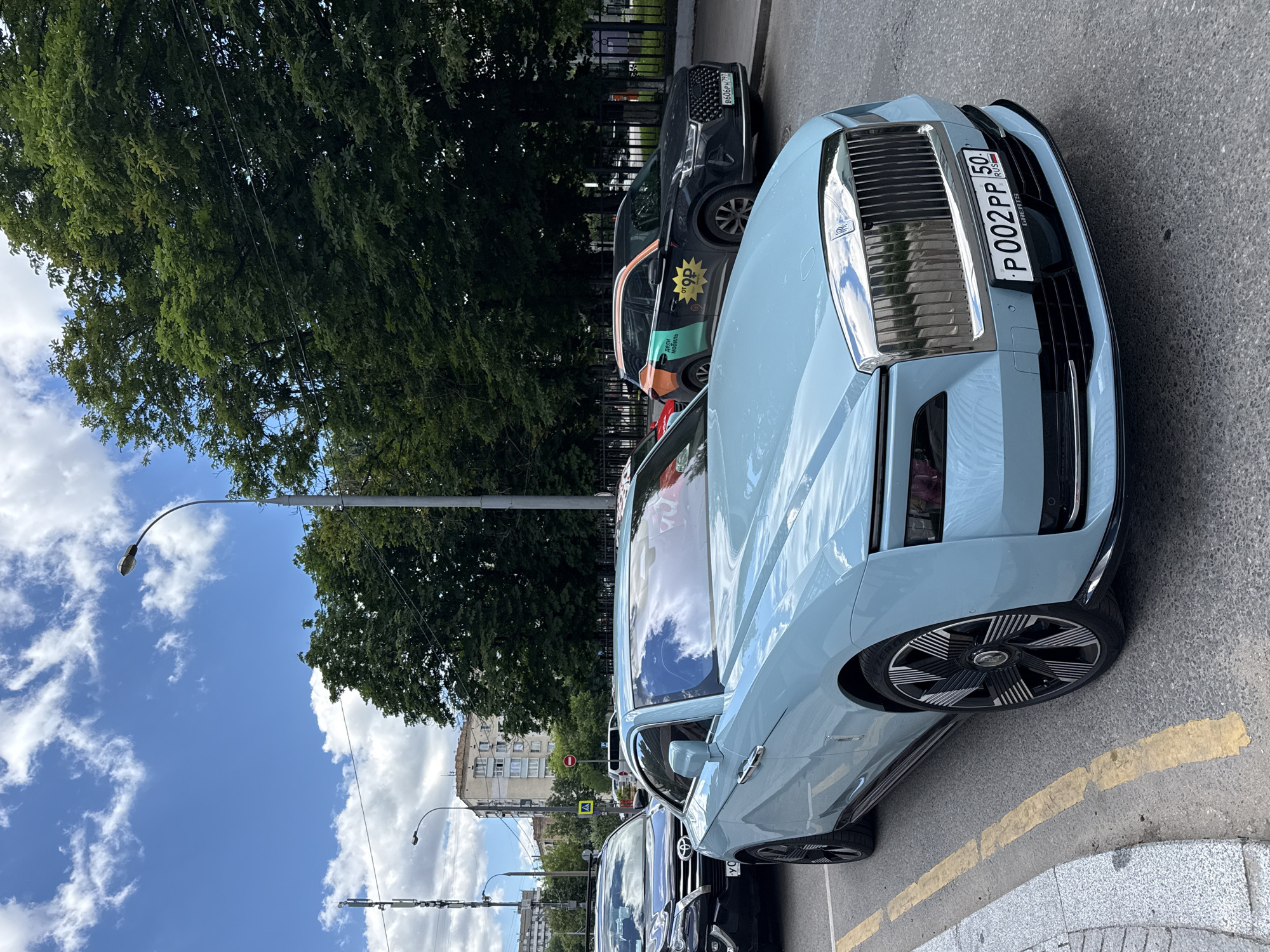 In Moscow, Russia, a pale blue Rolls-Royce coupe is parked on a street. The car's license plate reads "P002PP 50 RUS". Behind it, a black SUV with colorful branding on its side is also parked. The street is lined with trees, and a modern building can be seen in the background. The sky is partly cloudy.