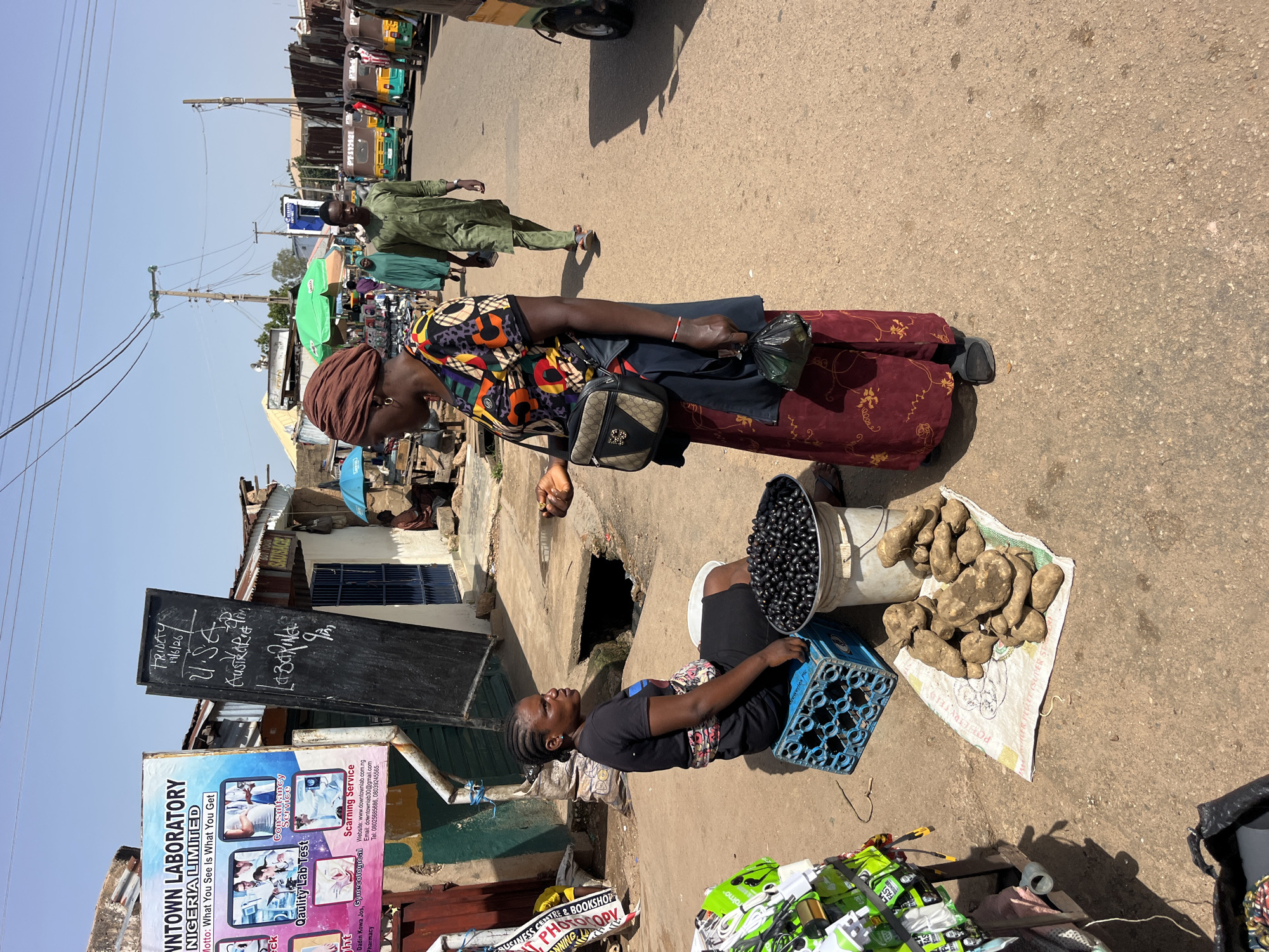 This media file depicts a street scene, likely a market, in an outdoor setting under a clear sky, suggesting daytime with fair weather. The perspective is from above, looking down at the ground and the individuals and objects present.
In the foreground, two women are the primary subjects. The woman on the right is seated on a concrete ledge, with her legs tucked beneath her. She is wearing a black t-shirt and a patterned skirt. Beside her, a blue plastic crate is visible, and to her left, a large white bucket is filled with dark, round fruits, possibly berries or olives. To the right of the crate, a pile of large, earthy-toned tubers, likely potatoes or yams, are spread out on a patterned cloth on the ground. The woman on the left is standing and is dressed in a colorful, patterned top and a long, dark red skirt. She is wearing a brown headscarf and has a small purse slung across her body. She is holding a black plastic bag.
Behind the women, the scene unfolds into a busy street. Several other people are walking in the background, some dressed in traditional attire. Various stalls or shops line the street, with some displaying goods and others appearing to be part of buildings. There are a few tricycles, commonly used for transport in some regions, visible further down the road. Electrical poles and wires are prominent features of the sky.
A significant detail is the presence of several signs with visible text. One is a blackboard with handwritten notes, including dates and place names like "FRIDAY 19/6/26", "U.S.A.", "Austria", and "LABORING 9 pm". Below this, a large, colorful advertisement poster for "DOWNTOWN LABORATORY NIGERIA LIMITED" is displayed, detailing their services and contact information, including "Consultancy Service", "Quality Lab Test", and "Scanning Service", along with a website and phone numbers. Another sign is partially visible with the word "SAUSAGE" on it. These textual elements provide crucial context, suggesting the location is Nigeria and potentially indicating a commercial or service-oriented area.
The overall impression is one of a bustling daily life in a developing urban or semi-urban environment, where commerce and social interaction are actively taking place. The presence of both produce and a laboratory advertisement hints at the diverse nature of the economic activities.