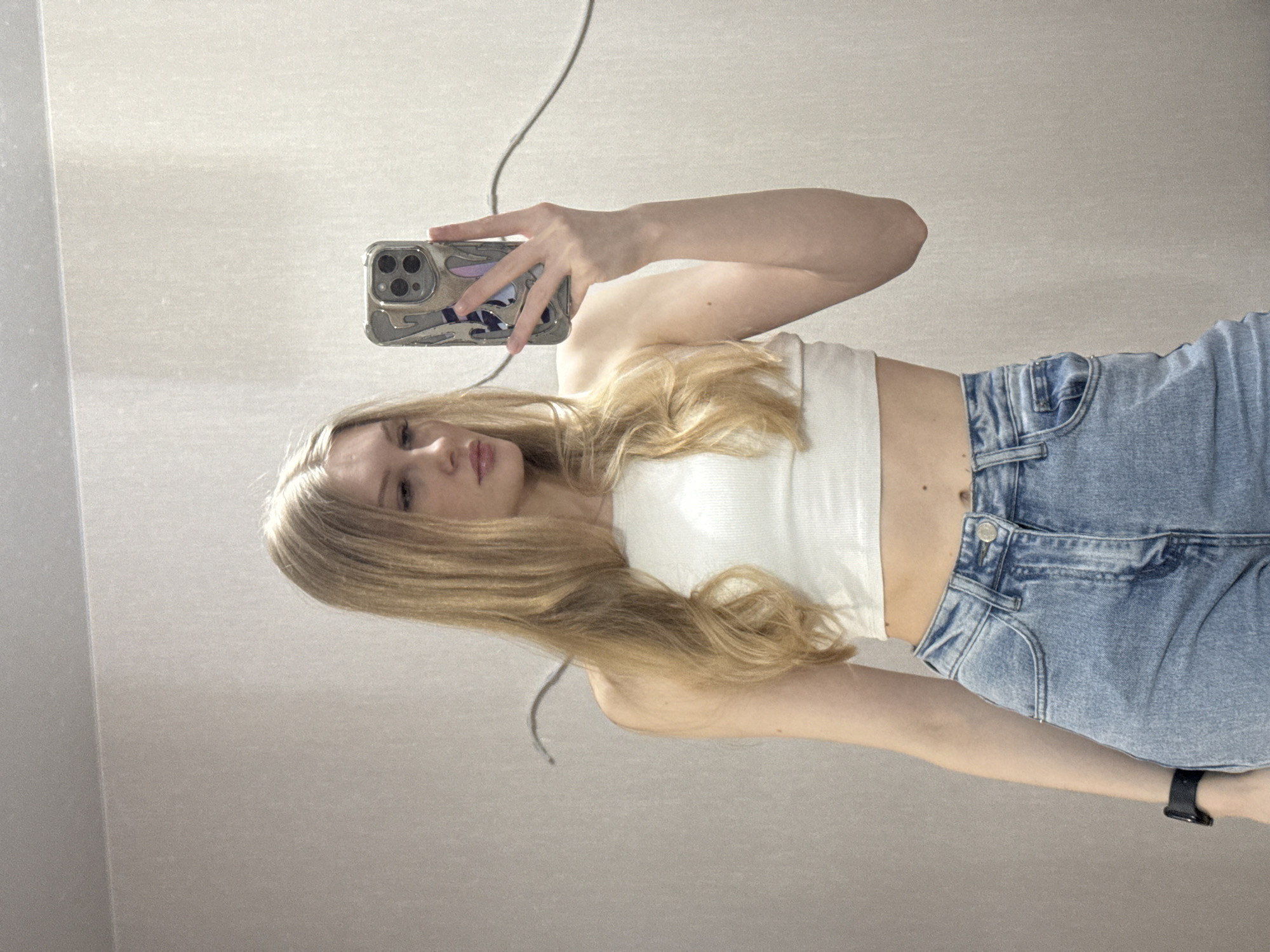 A young woman is captured in a mirror selfie, holding a smartphone with a distinctive patterned case in her right hand. She has long, straight blonde hair that cascades over her shoulders and chest. She is wearing a white, ribbed strapless crop top that reveals her midriff and high-waisted, light blue denim jeans. A black smartwatch or fitness tracker is visible on her left wrist.
The woman has a neutral to slightly pensive expression, looking directly towards her reflection in the mirror. The background consists of a plain, light-colored wall or surface, possibly off-white or light gray, suggesting an indoor setting. There are subtle textures and reflections on the wall surface. A thin, dark cable or string hangs partially visible in the upper left of the frame. The lighting is bright and even, indicating daytime. While the external location context is provided as Perm, Russia, there are no specific visual cues within the image to confirm this location. No discernible text is visible on her clothing or the objects, though the phone case has an intricate, raised design. The overall impression is a casual, contemporary portrait.