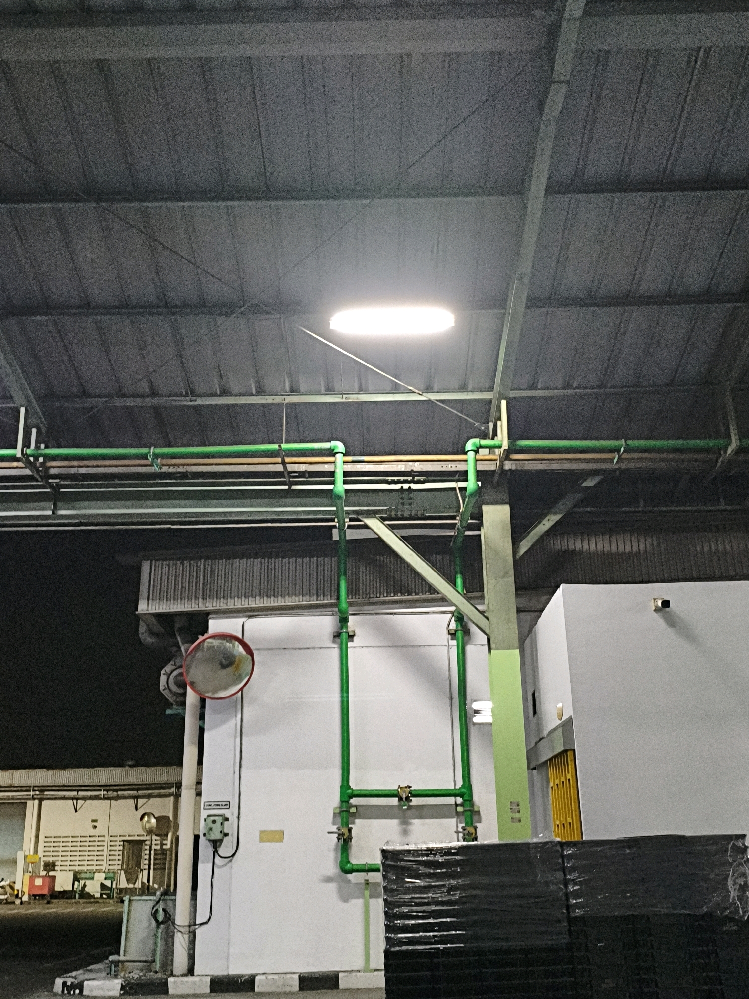 The image captures an interior view of a building, likely a warehouse or industrial facility, in Karawang, Indonesia. The scene is illuminated by a bright, linear fluorescent light fixture suspended from the corrugated metal ceiling. A network of green pipes, some supported by metal brackets and others by lighter colored pipes, runs horizontally and vertically throughout the space.
On the left side of the image, a large, round convex mirror with a red frame is mounted on a white wall. Below the mirror, a grey electrical box is attached to the wall, with a black cable running downwards. Further left, a section of a building with white louvred windows is visible, with some red and yellow equipment in the foreground, suggesting activity or storage.
The right side of the image shows a large, plain white wall, with a small black security camera mounted on it. In front of this wall, stacked black materials, tightly wrapped in plastic, occupy the lower portion of the frame. A bright yellow folding door is partially visible on the far right. The overall environment appears to be clean and functional, typical of an industrial or storage setting.
