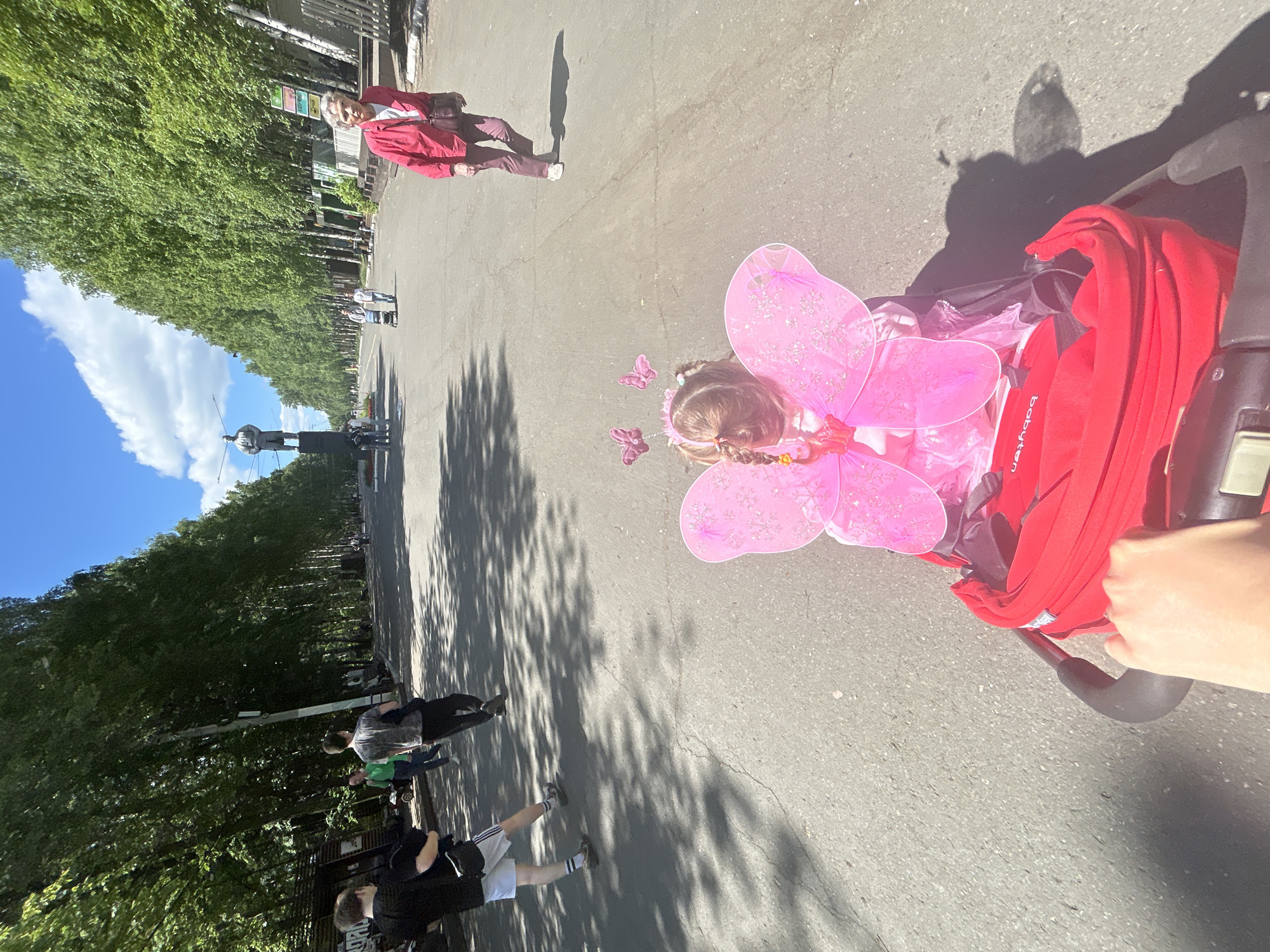A sunlit outdoor scene captures a path lined with lush green trees and various pedestrians. The image is taken from a slightly elevated perspective, looking down and along a wide avenue or park pathway.
In the foreground, a young child is seated in a red stroller, dressed in a vibrant pink fairy costume with large, glittery pink wings and small pink butterfly hair accessories. Part of a hand is visible pushing the stroller, indicating an adult is accompanying the child. The brand "babyten" is visible on the side of the red stroller.
Further along the path, an older woman in a red jacket, dark pants, and white shoes walks towards the viewer. Several other people are scattered in the mid-ground and background, walking or standing.
The path is paved, and strong, elongated shadows of trees and people stretch across it, suggesting a sunny day. The sky above is bright blue with scattered white clouds. In the far distance, a prominent large dark statue of a standing figure on a pedestal is visible, situated between the rows of trees. This statue, combined with the location context, identifies the scene as Izhevsk, Russia, likely depicting the Monument to Kuzebay Gerd.
The overall atmosphere is one of a pleasant, leisurely outdoor outing on a clear, bright day.