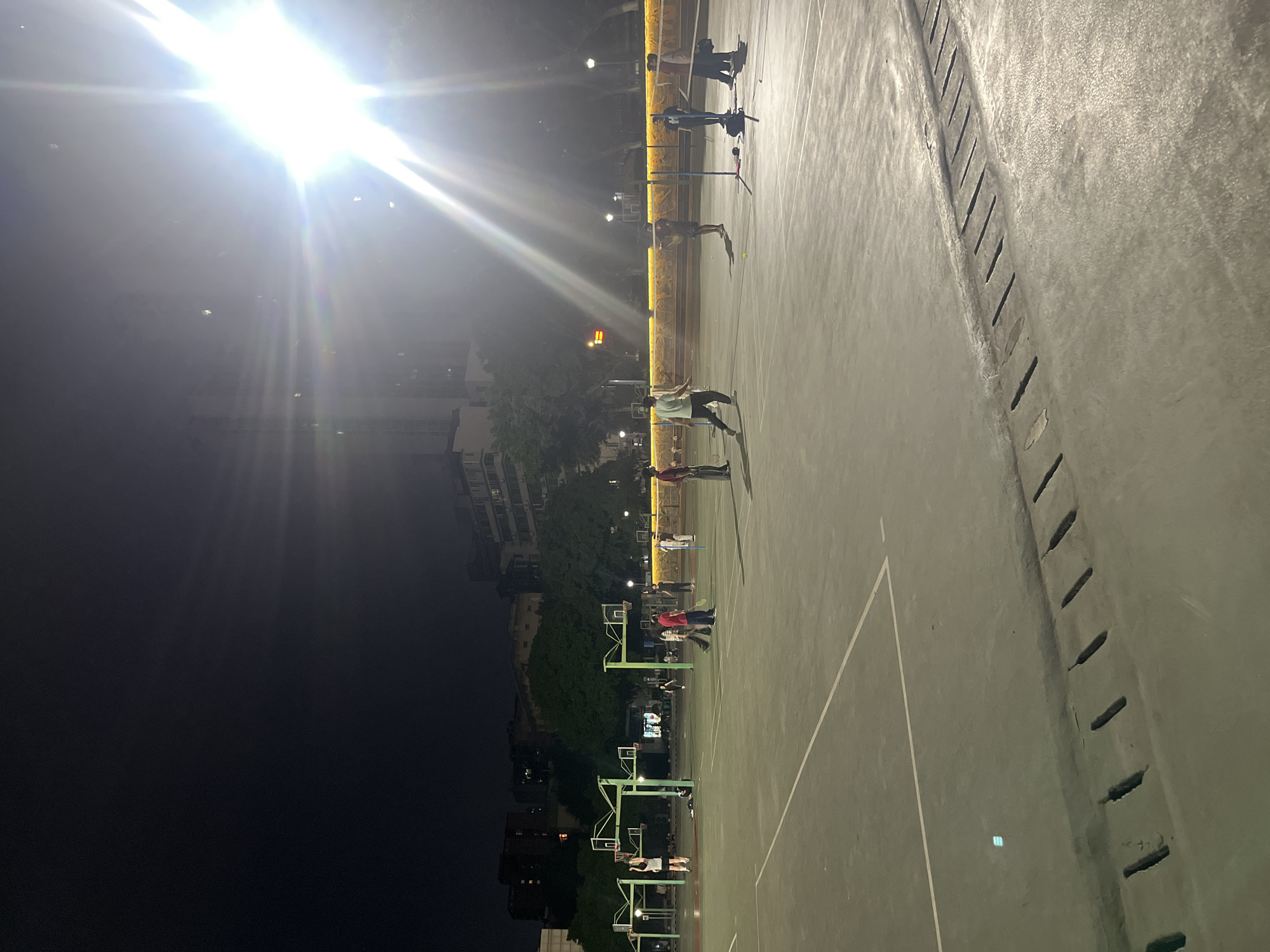 A long-angle outdoor night shot depicts a brightly lit, empty basketball court in Yuzhong District, China. People are playing badminton with rackets and shuttlecocks. A bright light source casts stark beams across the frame. Basketball hoops are visible. Buildings are in the background. A concrete drainage ditch borders the court.