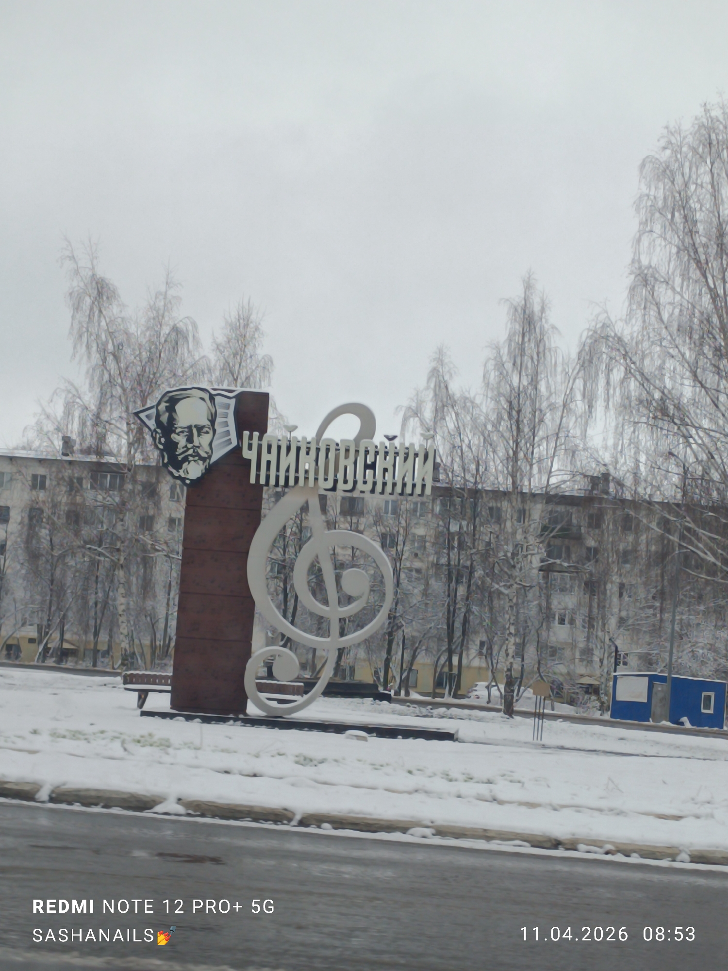 The image shows a winter scene in Chaikovsky, Russia. A monument dedicated to the composer Pyotr Ilyich Tchaikovsky is the main subject. The monument features a portrait of Tchaikovsky on a brown vertical slab, adjacent to a stylized white treble clef and the Cyrillic spelling of "Tchaikovsky." The ground is covered in snow. Bare trees stand in the background, partially obscuring a multi-story apartment building. A bench is visible behind the monument. A blue booth is positioned to the right. The sky is overcast. The bottom of the image displays text indicating the photo was taken with a Redmi Note 12 Pro+ 5G, a username, and a timestamp of 11.04.2026 08:53.