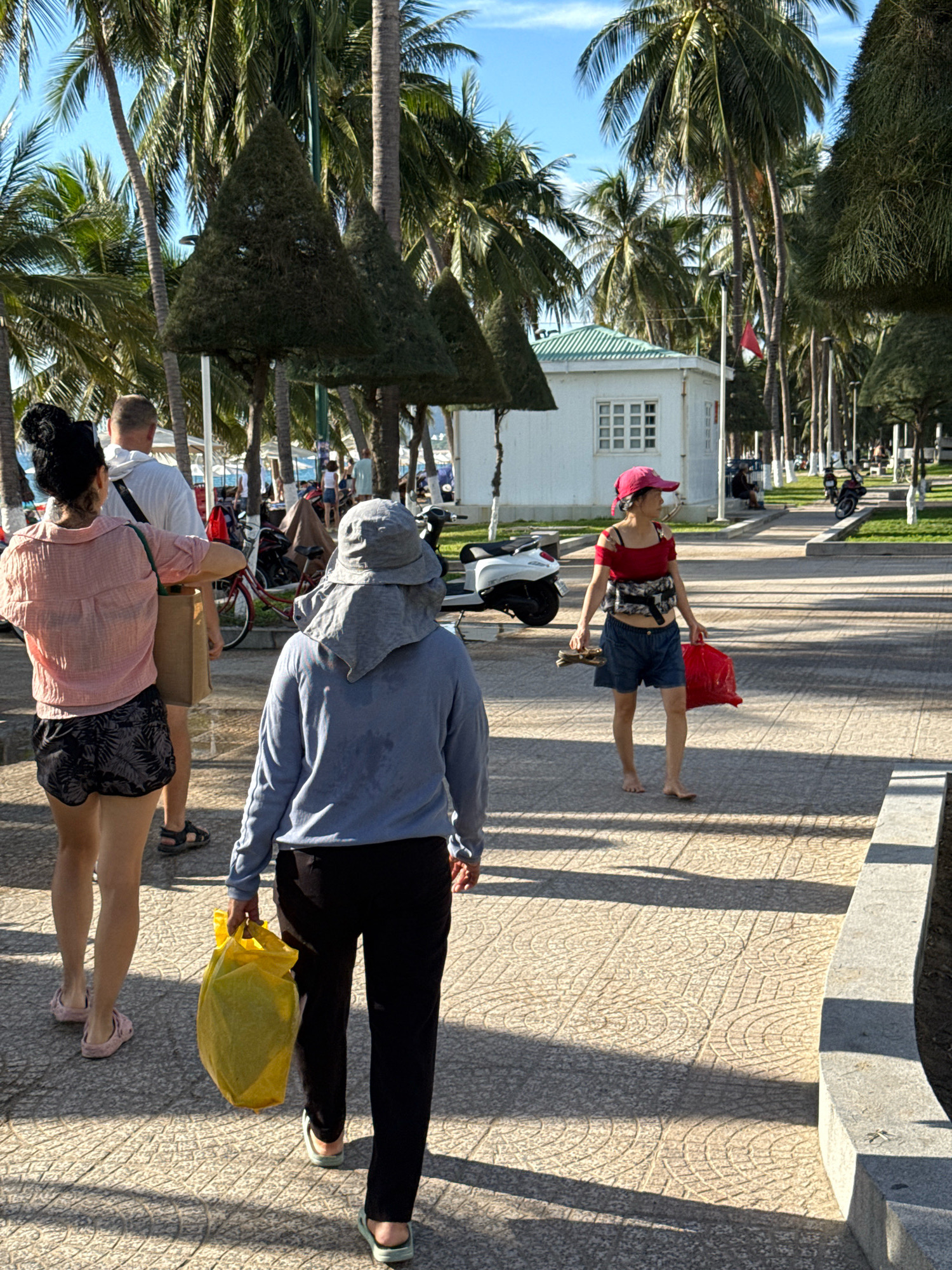 The image depicts a sun-drenched paved walkway lined with palm trees and stylized triangular hedges. In the foreground, three individuals are walking away from the camera. The person on the left is a woman with dark hair pulled back, wearing a pink patterned shirt and black patterned shorts. She is holding a brown tote bag over her shoulder. To her right is a man in a white hoodie and shorts, with a black strap across his chest. The third person, in the center, is wearing a blue long-sleeved shirt and black pants, carrying a large yellow plastic bag in their left hand. Further down the walkway, a fourth person, a woman wearing a red top, denim shorts, and a pink cap, is walking towards the camera, holding a red plastic bag and a pair of sandals. The environment suggests a coastal or resort area, likely in Nha Trang, Vietnam, given the prominent palm trees and the style of the architecture visible in the background, including a white building with a green roof. The ground is paved with patterned tiles, and shadows from the trees and people are cast across the walkway. In the distance, a red flag is visible on a pole, and several scooters are parked along the side of the path.