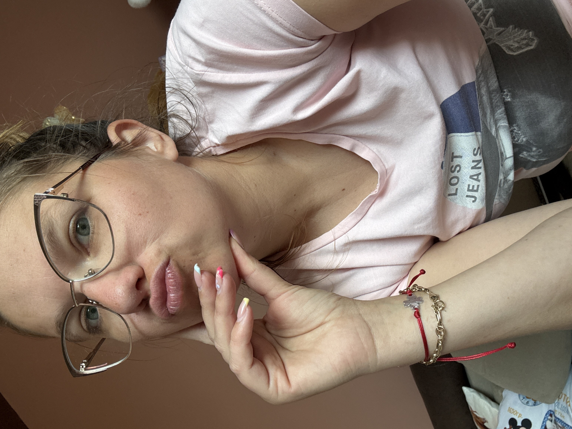 A woman is indoors, oriented at an angle to the camera, wearing glasses and a pink t-shirt with the text "LOST JEANS" visible. Her face is partially obscured by her hand, which is touching her cheek. She is wearing a bracelet consisting of a red cord and a gold chain. The background is a plain, light brown color. There is no information available about the location in Sofia, Bulgaria from the image.
