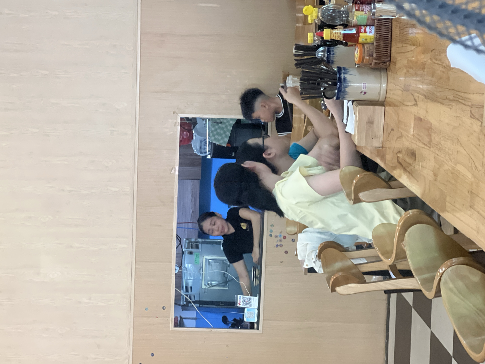 This is a medium shot photograph taken inside a restaurant in the city of Da Nang, Vietnam. The scene depicts a dining area with wooden tables and chairs, and a serving window in the background.
In the foreground, a light-colored wooden table is laden with various items. A white paper napkin dispenser sits to the left, next to a wooden box. A blue and white ceramic container filled with chopsticks and other utensils is prominently placed in the center, with "Hello Kitty" written on it. To the right of the utensil holder, a woven basket contains small containers, and several bottles of condiments are arranged, including what appears to be a bottle of chili sauce and other sauces with yellow caps. A glass bottle of water is also visible. The table has a visibly textured, polished wooden surface.
In the middle ground, several people are seated at the table. On the left, a person wearing a light yellow top is seen from the back, with their head turned to the right. Next to them, another person wearing glasses and a blue top is also seen from the side, facing towards the right. Further to the right, a young boy with a distinct haircut, wearing a black t-shirt with white trim, is looking to his left.
In the background, a serving window looks into the kitchen area. A woman in a black t-shirt is standing behind the counter, reaching out towards the left. She appears to be interacting with something or someone outside the frame. Behind her, kitchen equipment, including a large stainless steel appliance, is visible. On the counter, near the woman, there are two small bowls and what appears to be a QR code displayed on a stand. The wall behind the serving window is blue, contrasting with the wooden paneling of the dining area.
The flooring visible in the lower left corner is a checkered pattern of dark and light squares, suggestive of tiles. The walls of the dining area are paneled with light-colored wood, and the ceiling appears to be a pale color with a wood-grain texture. The overall lighting suggests an indoor setting, possibly during the daytime.