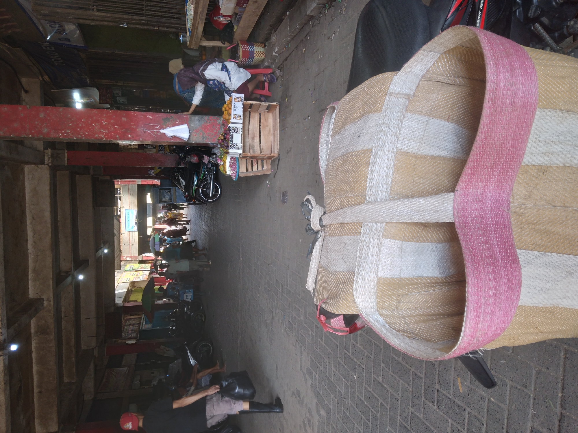 The image depicts a market scene in Purwokerto, Indonesia, focusing on the foreground where a large woven bag sits atop a motorcycle. The bag is constructed from light brown and white woven material, with a distinct pink woven band around its opening. In the background, the market is bustling with activity. Several people are visible walking through the market, some carrying items. Motorbikes are parked in various locations throughout the area. A woman wearing a hijab is seated on a stool next to a stall displaying fruits, which are arranged in crates. The market appears to be covered by a roof supported by pillars, one of which is prominently red and shows signs of wear. The ground is paved with interlocking bricks. The overall impression is of a vibrant, open-air marketplace.