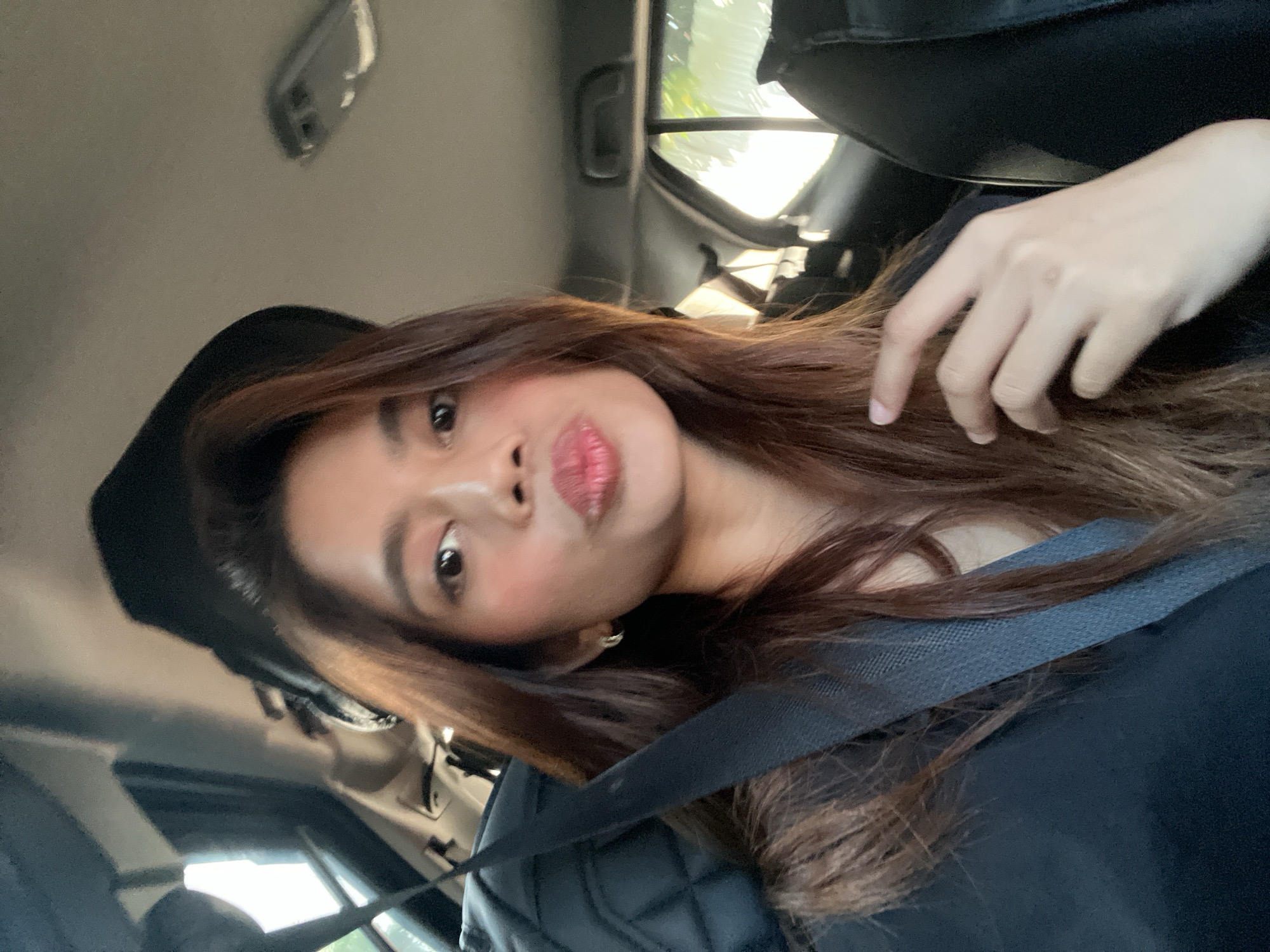 A young female, appearing to be in her twenties, is seated as a passenger inside a vehicle, capturing a selfie. She has long, reddish-brown hair, parted to her left, and is wearing a dark-colored top. A black seatbelt is visible across her chest and shoulder. Her lips are puckered in a "kiss" pose, and she has visible blush on her cheeks and light eye makeup. A silver hoop earring is worn on her left ear. Her left hand, with slender fingers, is resting gently on her chest, near her hair. The car's interior features a light grey or beige headliner and dark, possibly black, quilted upholstery on the seat to her left. Bright daylight streams through the windows, with blurred green foliage visible outside the rear passenger window, suggesting an outdoor, possibly urban or suburban, setting. The scene is located in Davao City, Philippines.
