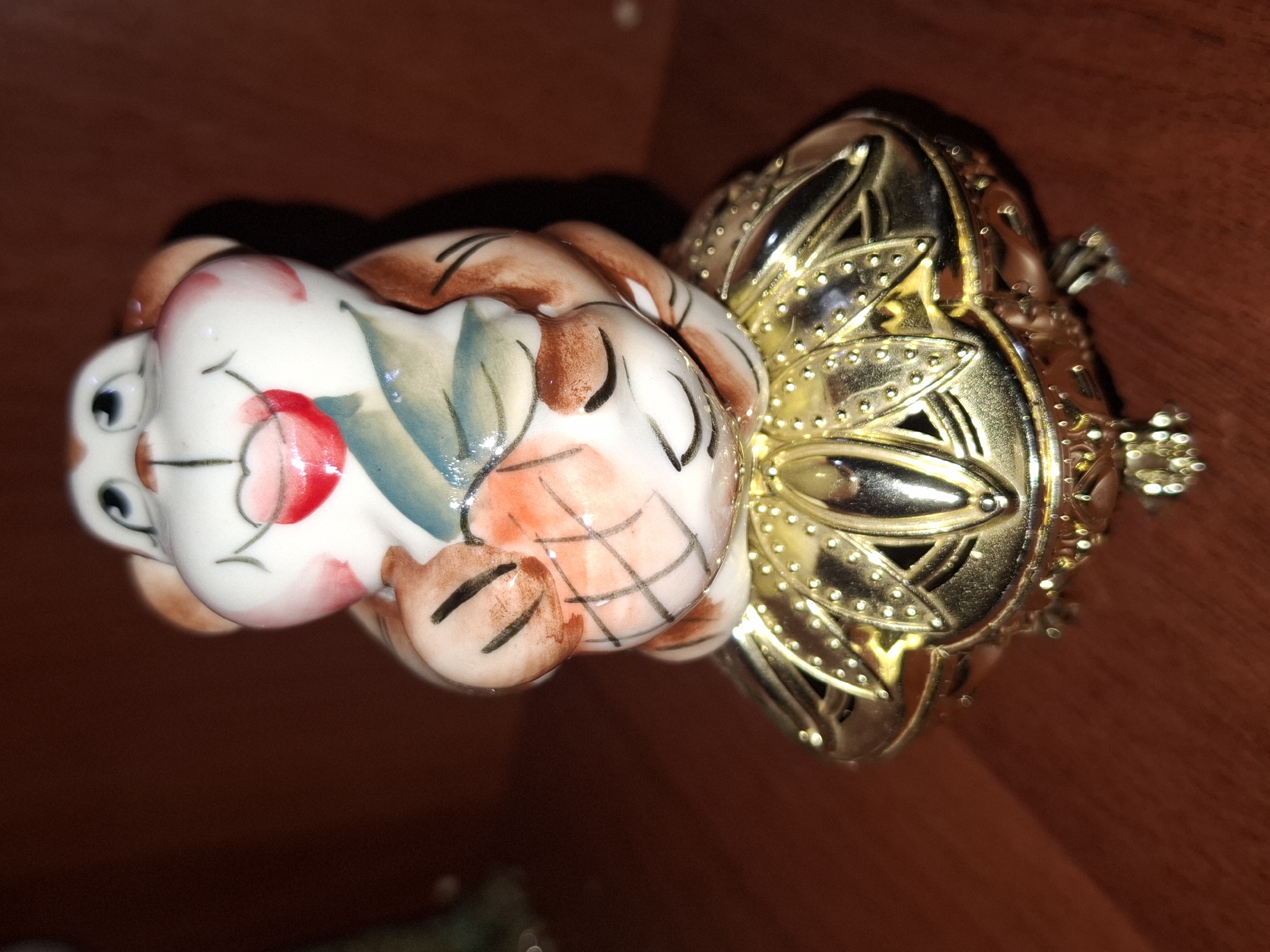 A ceramic figurine of a monkey sits atop a golden decorative box with intricate cut-out designs and small feet. The monkey is white with brown accents and a red mouth, holding what appears to be a fruit or vegetable. The figurine and box are placed on a polished wooden surface. The background is a blurred, dark brown color, likely a wall or shelf. The scene is indoors, likely a home environment. There is no indication of the city of Almaty, Kazakhstan.