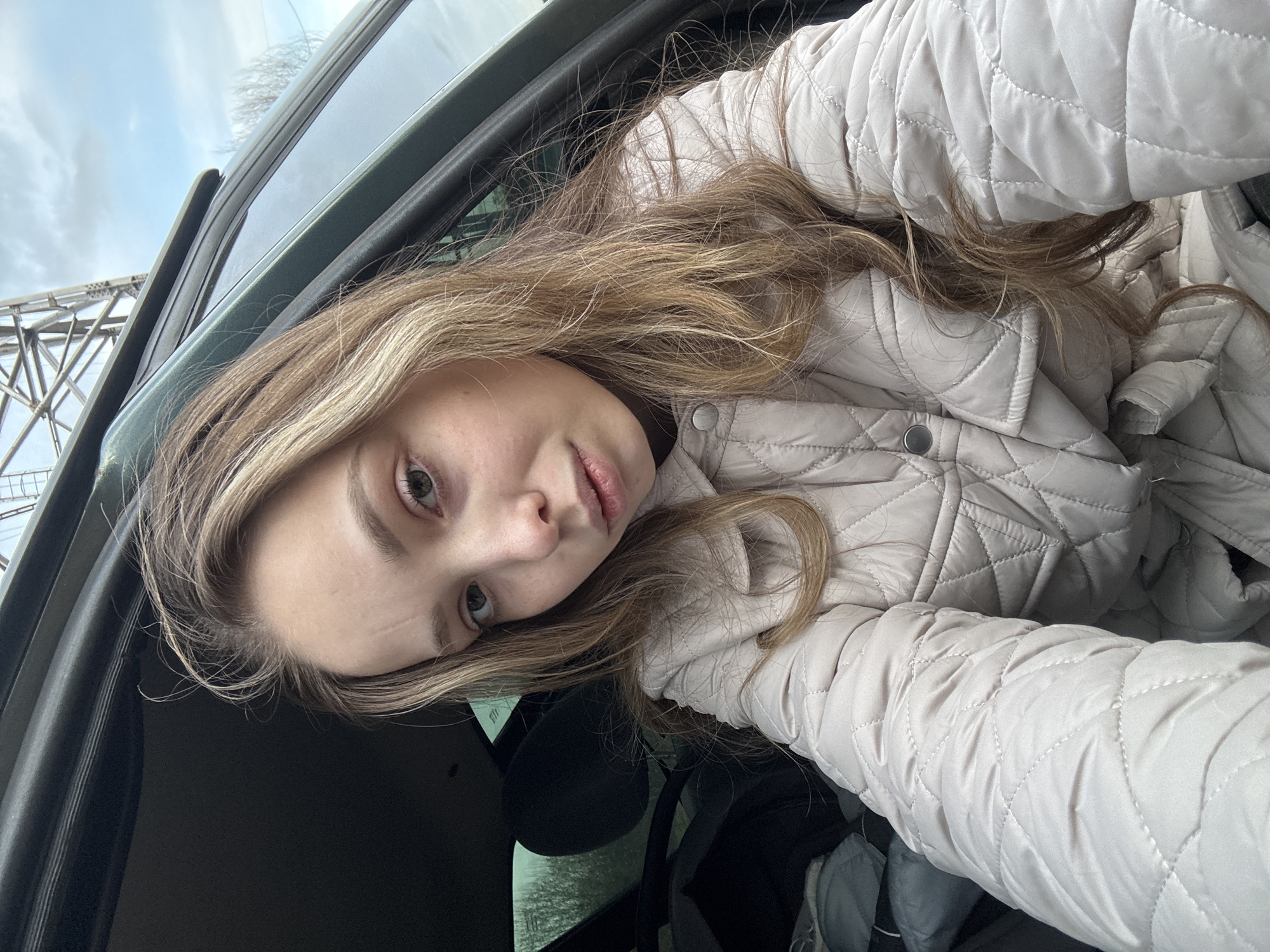 A young woman with long, light brown hair is taking a selfie inside a vehicle, possibly a car. She is wearing a light beige quilted jacket. The vehicle's window and frame are visible in the background, along with a glimpse of a bridge or similar structure. Based on the prompt, the location is the city of Chaikovsky, Russia.