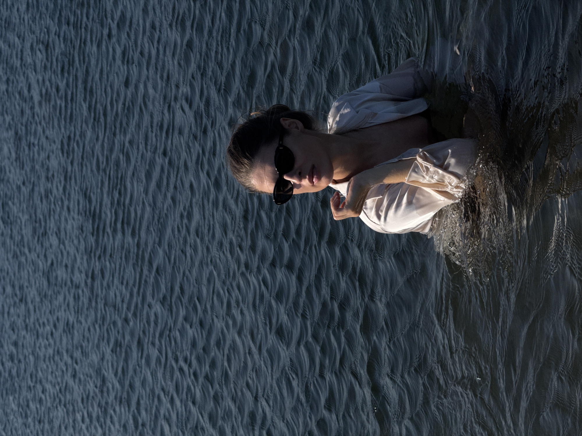 A woman wearing a light-colored satin blazer and black sunglasses stands in dark, rippling water. The blazer is unbuttoned, revealing a dark garment underneath. Her hair is pulled back, and she is looking to her left, with her left hand resting on her chin. The water is choppy, with small waves and some areas of splashing. The background consists entirely of the dark water. The scene is likely outdoors, given the natural lighting and water. The location is not specified within the image itself.