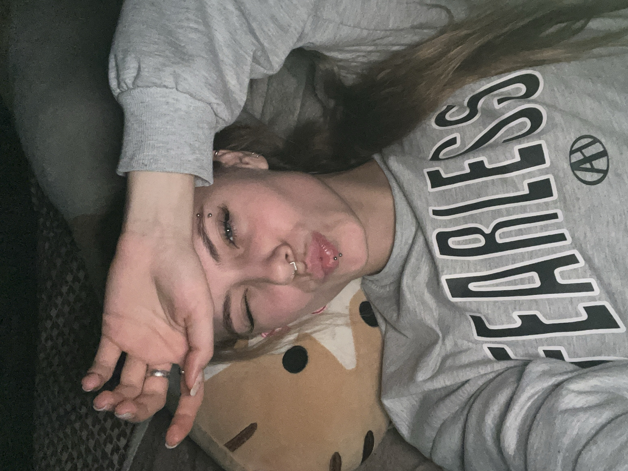 A young woman with multiple piercings (eyebrow, nose, lip, ear) is lying down, her face partially obscured by her hand. She is wearing a gray sweatshirt with the word "FEARLESS" printed in bold, black letters. Her head rests on a plush cushion featuring a brown and black pattern. The background is indistinct, suggesting an indoor setting with subdued lighting.
