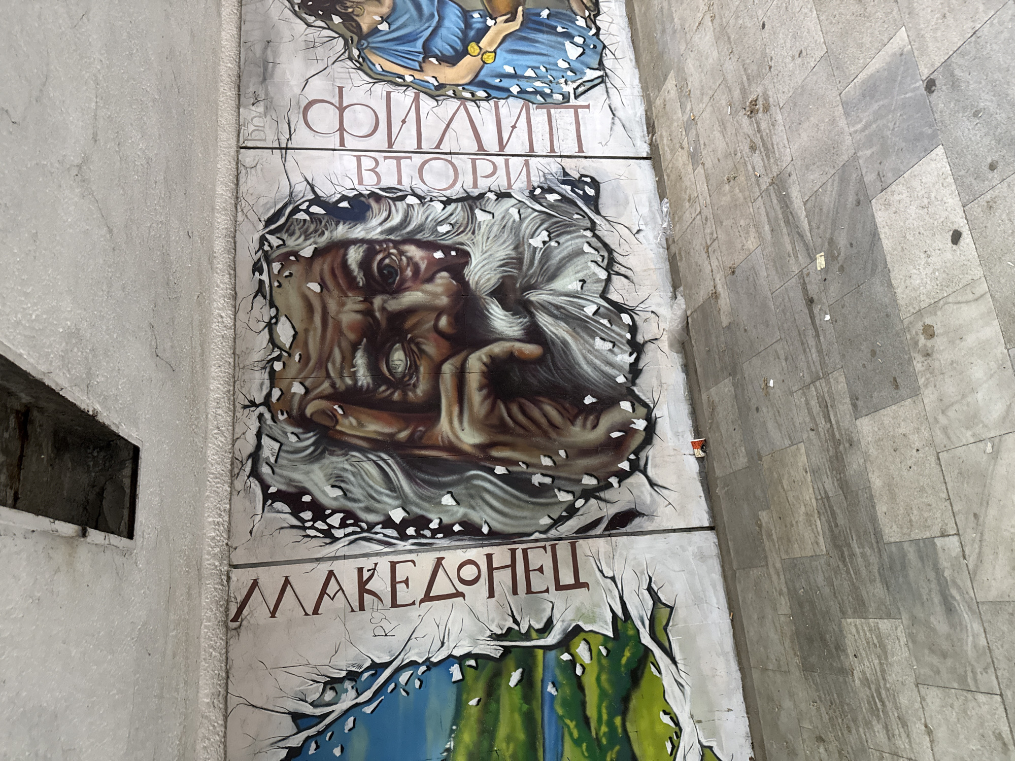 The image depicts a section of a large, weathered mural or public art installation on a light-colored wall, presumably outdoors. The mural is divided into at least three horizontal panels, each separated by apparent cracks or breaks in the surface. The topmost panel partially displays a figure in flowing blue garments. The second panel features a close-up portrait of a man with long, white hair and beard, appearing aged. His expression seems contemplative. The third panel visible reads "МАКЕДОНЕЦ" in a red-brown font, and shows abstract shapes of green and blue colors. The surrounding environment includes a light-colored wall with a rectangular opening and a stone-tiled area on the right. The location is likely Plovdiv, Bulgaria, given the Cyrillic script used in the writing on the mural.