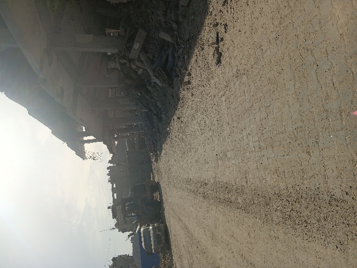 The image depicts a scene of extensive urban destruction in the city of Khan Yunis, Palestinian Territory. In the foreground, an unpaved dirt road with visible tire tracks and scattered debris dominates the lower half of the frame. To the right, a severely damaged multi-story concrete building stands with several upper floors partially collapsed, exposing reinforced concrete slabs and vertical support columns. A large mound of concrete rubble and shattered building materials is piled at the base of this structure, extending into the roadway. On the left side of the road, a white van is parked. Behind the van, a dark-colored heavy construction vehicle, likely a backhoe or excavator, is stationary. Further back, part of a larger utility or dump truck is visible. The background shows additional buildings, some appearing damaged, under a bright, hazy sky. No people are visible. The scene suggests the aftermath of significant structural damage or demolition.
