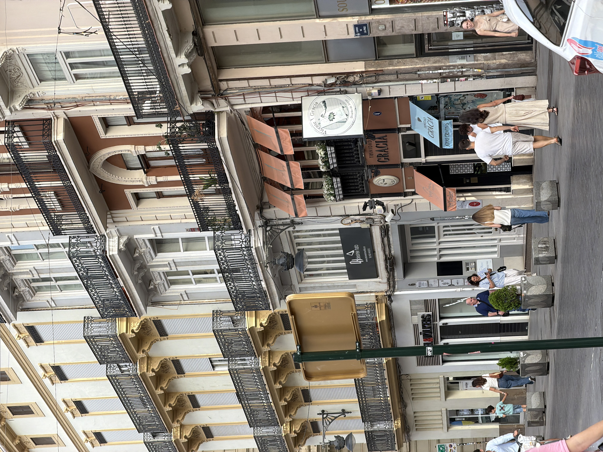 The image shows a street scene in a city, likely in Europe, with pedestrians walking past shops and ornate buildings. The architecture features balconies with black railings, decorative cornices, and multiple stories. Several storefronts are visible, including one with awnings and another with a sign that reads "TRINIDAD GRACIA JOYAS CREATIVAS." People are dressed in casual summer clothing. A green traffic pole with a yellow sign is prominent in the foreground. The overall impression is of a bustling urban environment.