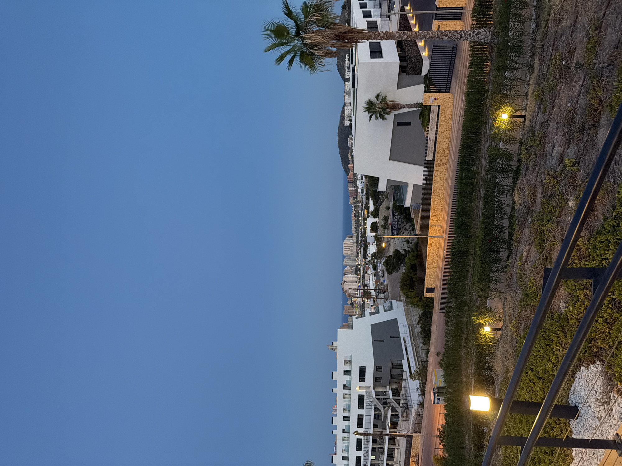 The image captures a twilight scene in Finestrat, Spain, overlooking a residential area with modern buildings and a distant cityscape. In the foreground, a black metal railing runs horizontally across the bottom, with white gravel and green ground cover beneath. Several low-lying outdoor lights with warm yellow illumination are visible along a path.
Further back, a series of low hedges and dry brush form a landscaped embankment. Beyond the embankment, a light-colored wall topped with a dark metal fence encloses a property. Two distinct modern buildings are prominent: one on the left is a multi-story white and gray structure with visible balconies and staircases, while the building on the right is a lower, more angular white structure with dark-framed windows. A tall palm tree stands to the right of this building.
In the mid-ground, a street with streetlights is visible, with some construction materials or equipment present. The background features a more densely built-up area with numerous high-rise buildings, likely the coastline or a larger town, situated by the sea. Rolling hills are visible behind the buildings. The sky is a clear, deep blue, indicative of dusk. The overall impression is of a contemporary development in a coastal Spanish location.