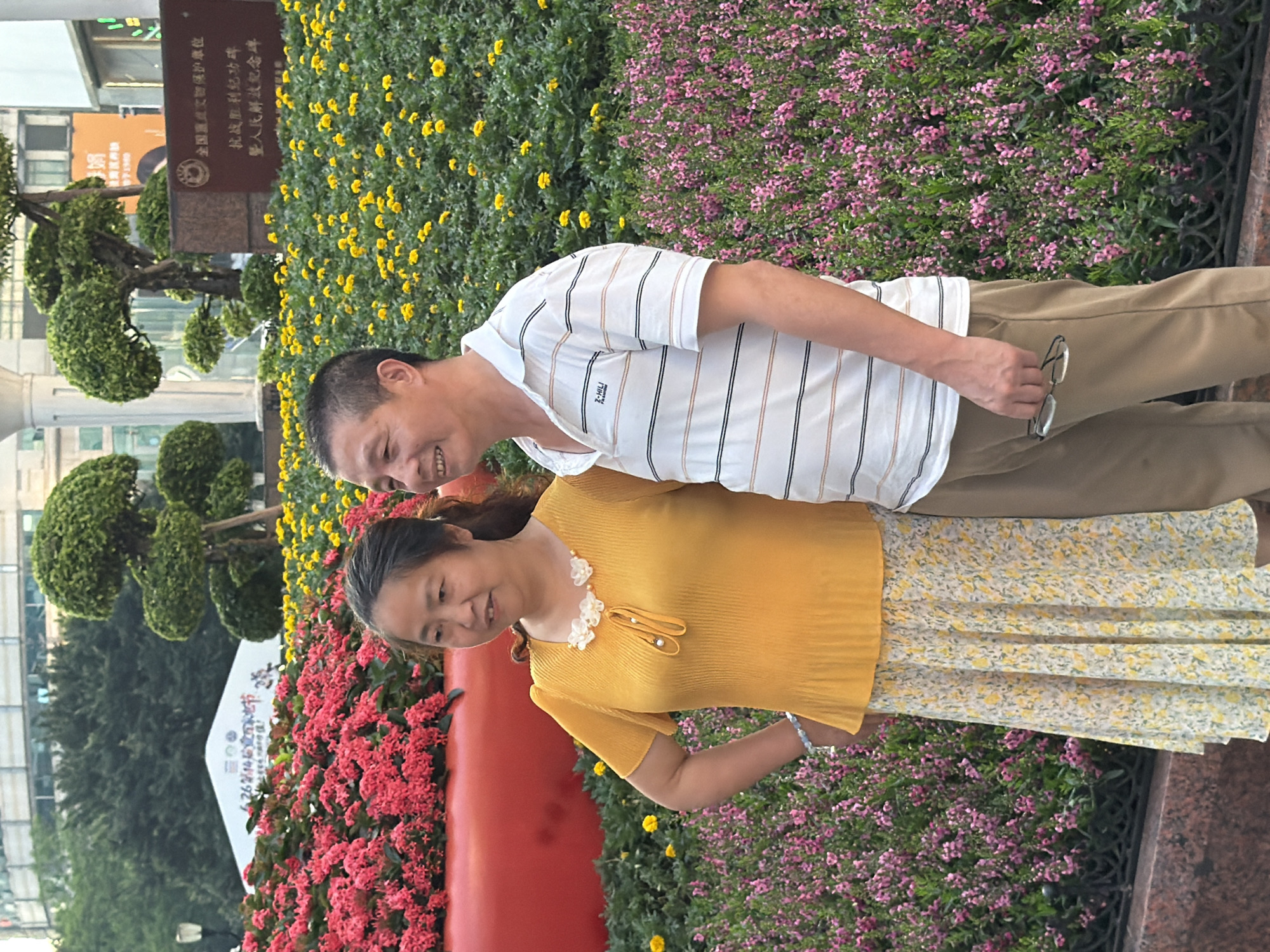 A middle-aged Asian couple stands in front of a floral display in what appears to be a public square or park in Yuzhong District, China. The woman, on the left, wears a bright yellow, short-sleeved, ruffled blouse and a yellow floral-patterned skirt. She has dark hair pulled back and is adorned with a beaded necklace and a bracelet. Her left hand rests on her hip. The man, on the right, is dressed in a white, striped polo shirt and khaki pants. He is holding a pair of glasses in his right hand. Both individuals are smiling and looking towards the viewer.
The background features a vibrant landscape of flowers. A large planter filled with bright red flowers dominates the left side of the frame, partially obscuring a white tent with text and graphics. Behind the couple and the red flowers, beds of small yellow flowers and pink flowers are visible. In the distance, there are trees, buildings, and what appears to be a monument or plaque with Chinese inscriptions. The scene suggests a well-maintained outdoor public space, possibly decorated for an event. The lighting indicates it is daytime, likely late afternoon or early evening due to the warmth of the light.