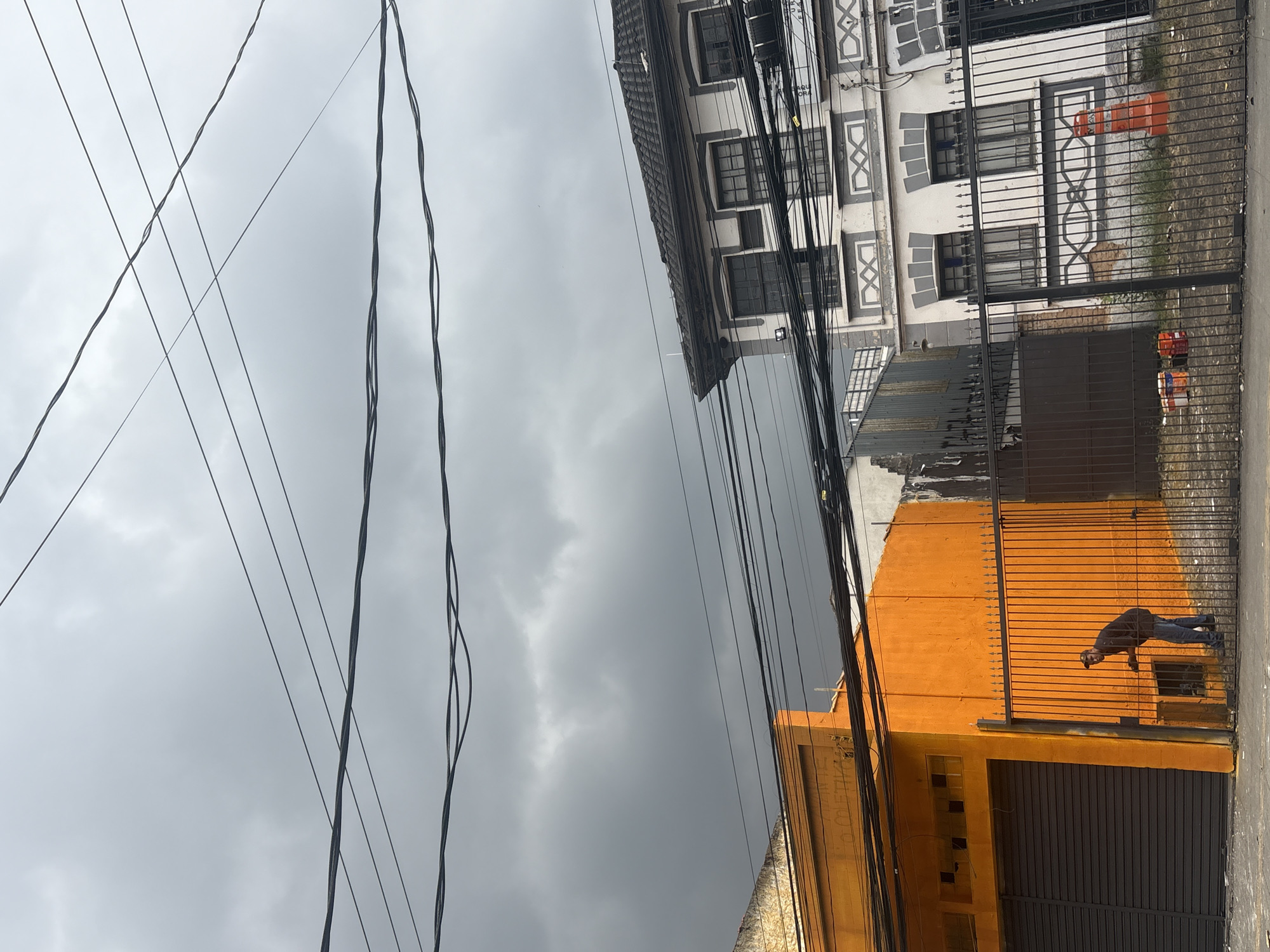 The image depicts a street scene in São Paulo, Brazil, under a heavily overcast sky with dark, ominous clouds. In the foreground, a black metal fence runs horizontally across the frame. To the left, a vibrant orange building with a dark gray metal rolling door is visible. A man wearing a dark jacket, blue jeans, and a baseball cap is standing near the fence, leaning towards the orange building, with his hands near an opening in the wall. He appears to be interacting with something within the opening. To the right of the orange building, a portion of a white building with gray decorative trim and multiple windows is seen. The building has a tiled roof and appears to be two stories high. More electrical wires crisscross the sky above both buildings. Behind the fence, on the ground, there are two paint buckets, one orange and one red, and an orange traffic cone. The overall impression is a somewhat gritty urban environment on a day threatening rain.