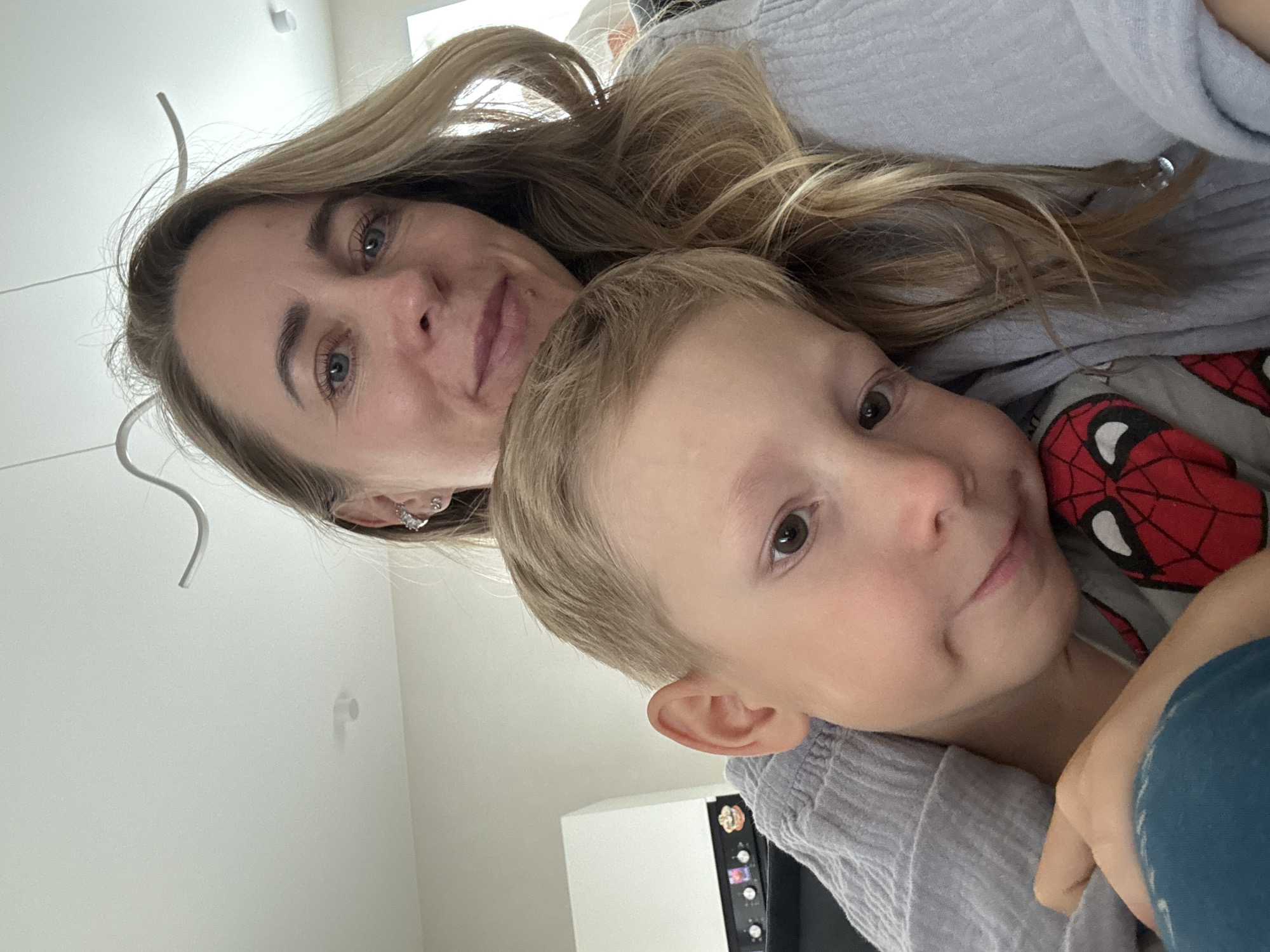 A woman and a young boy are close together in what appears to be a bedroom. The woman is on the left, with blonde hair and blue eyes, wearing a grey sweater. The boy is on the right, with blonde hair and brown eyes, wearing a shirt with a Spiderman design. They are both looking towards the camera, and both have gentle smiles. The scene is brightly lit, suggesting it is daytime. The background is blurred but shows hints of furniture, possibly a dresser or cabinet. The overall mood is one of affection and comfort.
