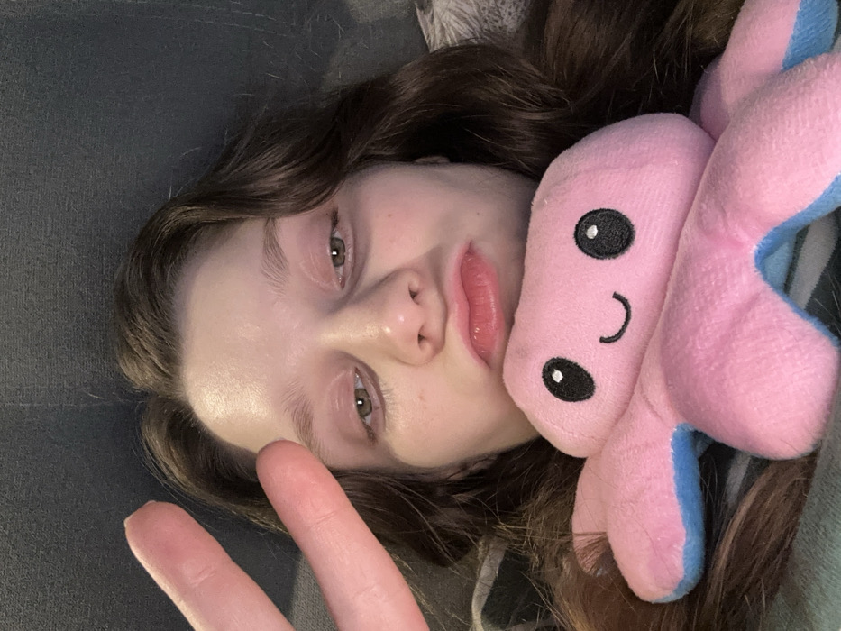 A young woman with long brown hair rests on a gray surface, likely a couch. She has fair skin and green eyes, and her lips are slightly pursed. A pink and blue reversible octopus plush toy rests next to her head. Her hand is visible, displaying a peace sign. The photo is taken at a tilted angle, with the subject and octopus lying down.