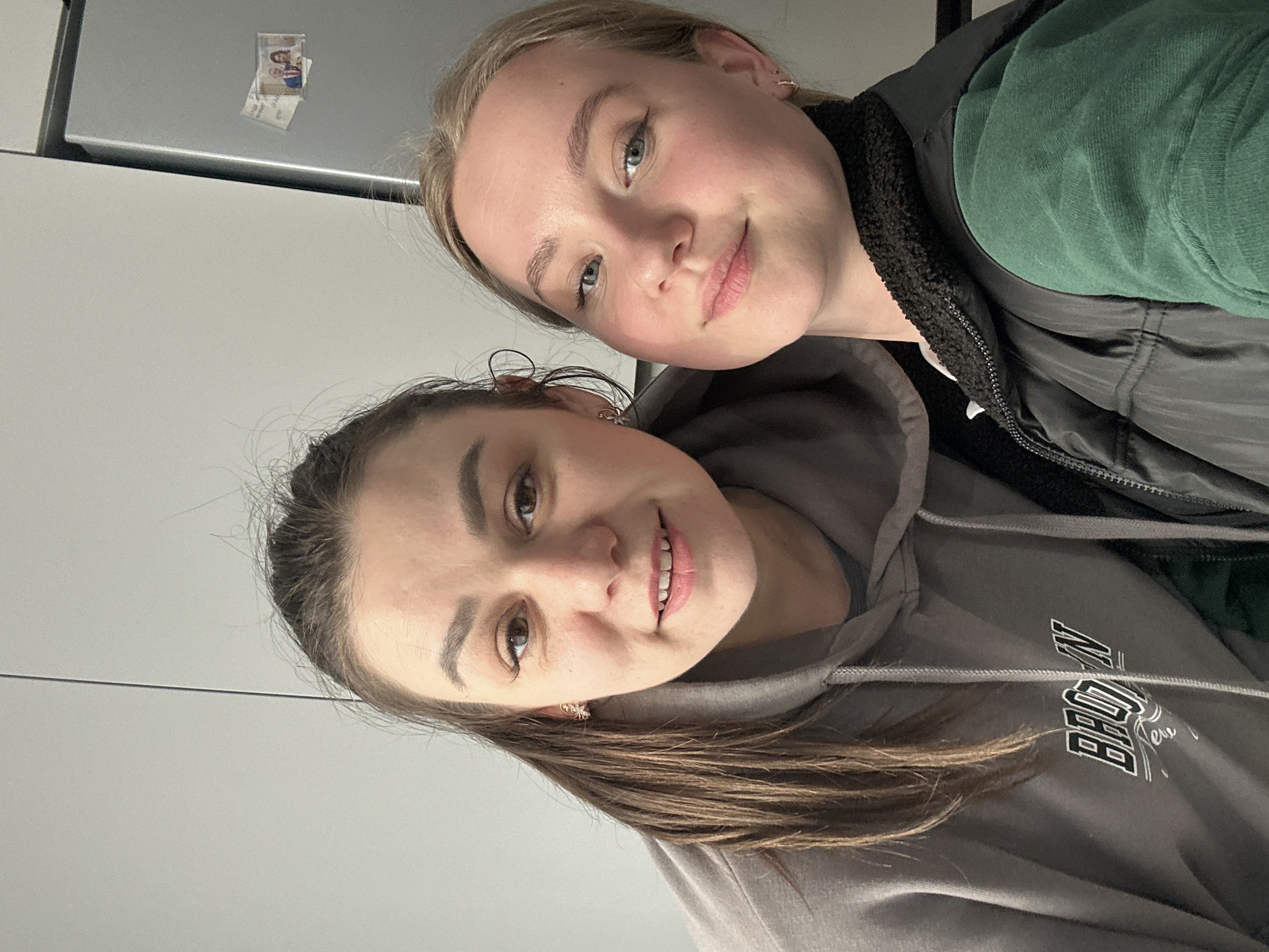 Two young women are captured in a casual, close-up indoor selfie, both smiling at the camera.
In the foreground, a woman with medium-brown hair, styled down, has dark eyes and is smiling. She is wearing a grey hooded sweatshirt with the word "BROOKLYN" partially visible in white text on the front. She has small, light-colored earrings.
Behind her, slightly to the right, another woman with blonde hair and blue-green eyes also smiles. She is wearing a dark green top layered under a black, possibly fleece-lined, vest. Small hoop earrings are visible in her ears.
The background appears to be an interior setting, possibly a kitchen or living area, with a light grey wall and a metallic surface visible behind the blonde woman's head. A small, framed photo, likely a magnet, depicting two individuals, is attached to this metallic surface. The overall mood is cheerful and relaxed.