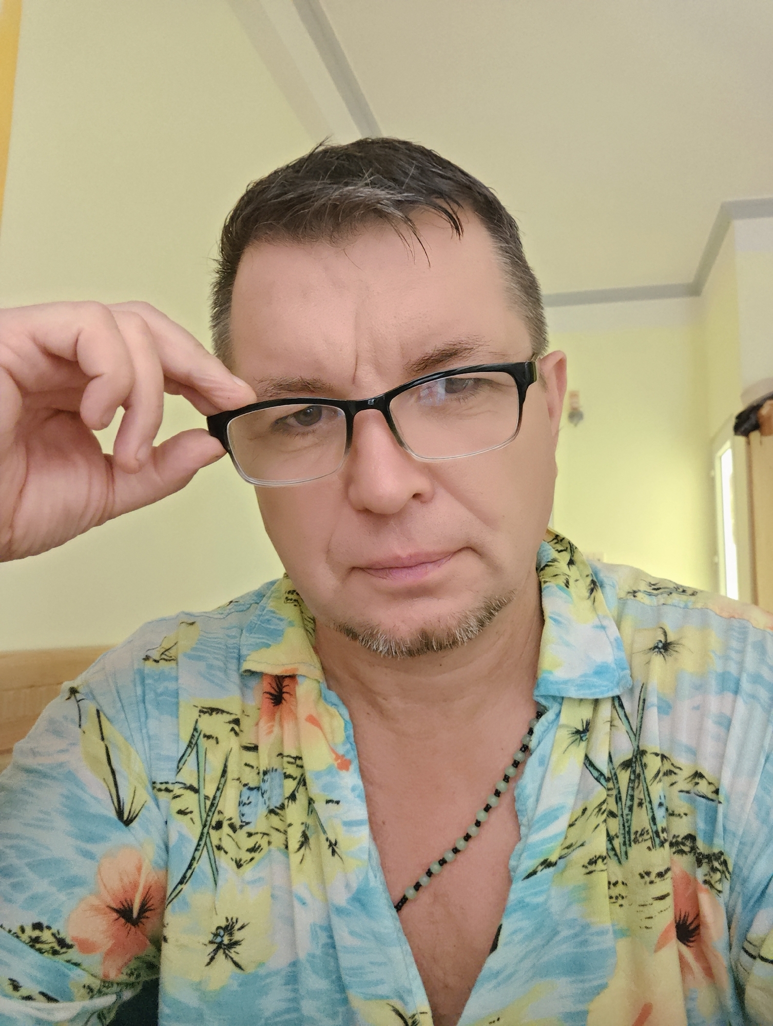 A medium shot photograph shows a middle-aged Caucasian male wearing glasses and a Hawaiian-style shirt. The man is looking directly at the camera, with his left hand on the temple of his glasses, adjusting them. He has short, dark hair with some graying, a goatee, and appears to be in an indoor setting. The background is a plain, light yellow wall with a gray trim at the top, suggesting an interior space. The man is also wearing a beaded necklace. The photograph is taken from the chest up, with the man centered in the frame. There is no visual information in the image that can be used to determine the location in Nha Trang Ward, Vietnam.