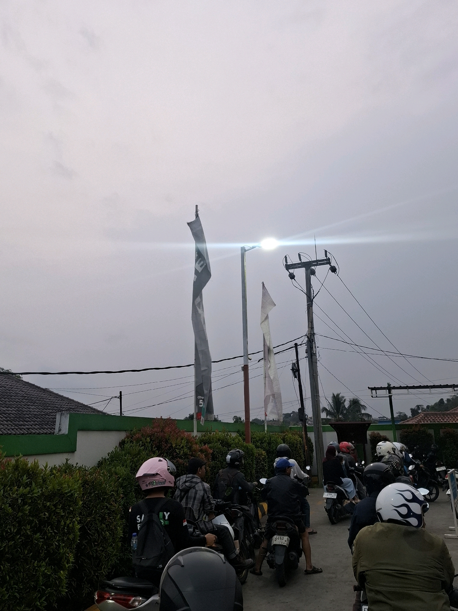 The image shows a street scene in Sukabumi, Indonesia. Several people are on motorcycles, some waiting in line and others moving. The sky is overcast, and there is a bright light illuminating the scene, possibly from a street lamp. There are also utility poles with wires, flags on poles, and buildings in the background. Green foliage lines the side of the road.