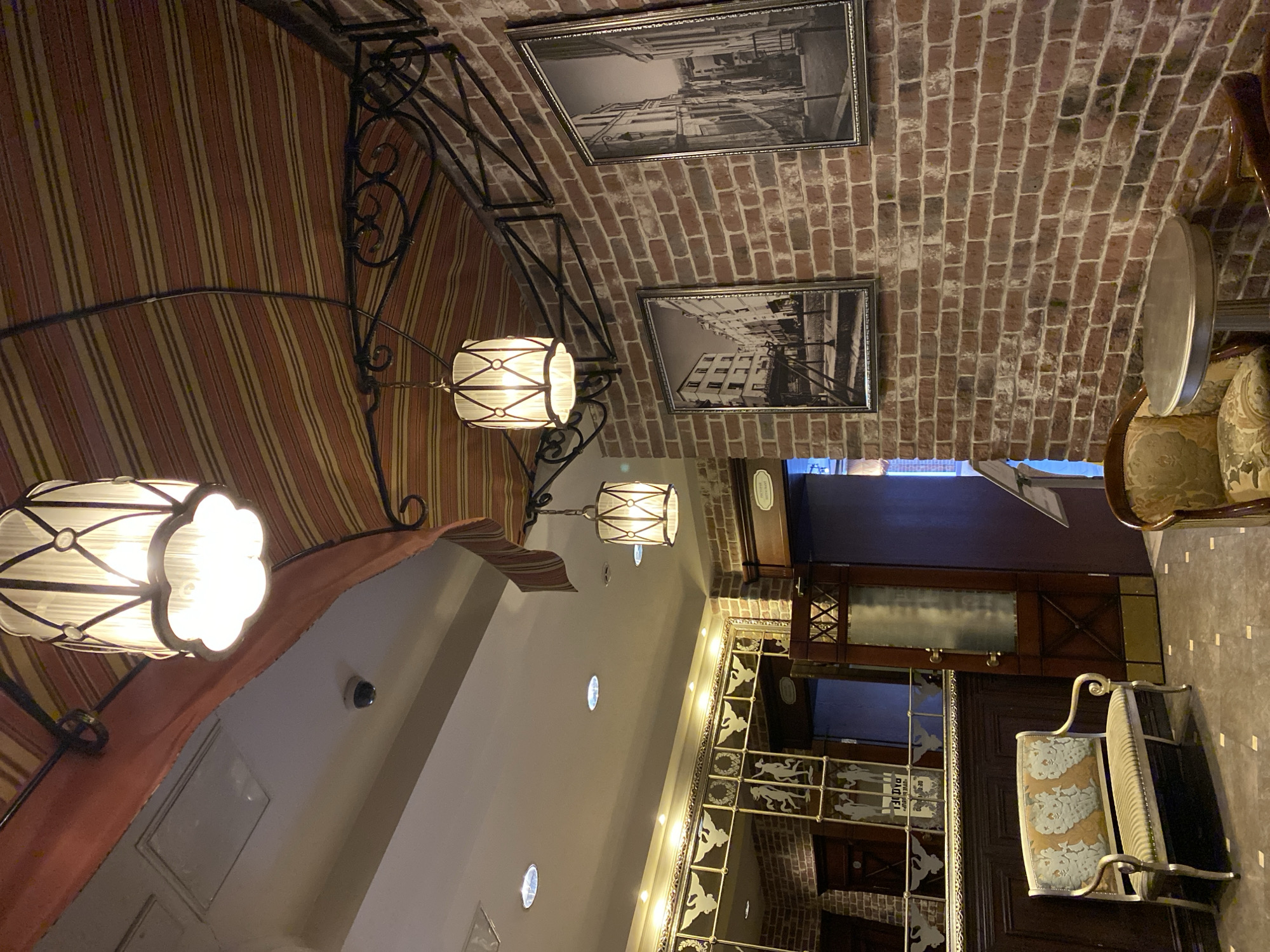The image depicts the interior of a restaurant. The ceiling is constructed from brick with two framed black and white photographs hung upon it. Below the brick ceiling, there is an elaborate ironwork structure that supports several hanging pendant lamps. A red and tan striped fabric covers a portion of the ceiling. The wall beneath is painted white, and there are several circular recessed lights. The other side of the image shows a dark wood-paneled wall, where an ornate screen is mounted. A upholstered chair with decorative metal legs is visible in the foreground. Given the restaurant's interior design, which features brickwork and vintage photographs, it is likely located in a historic building in Yekaterinburg, Russia.