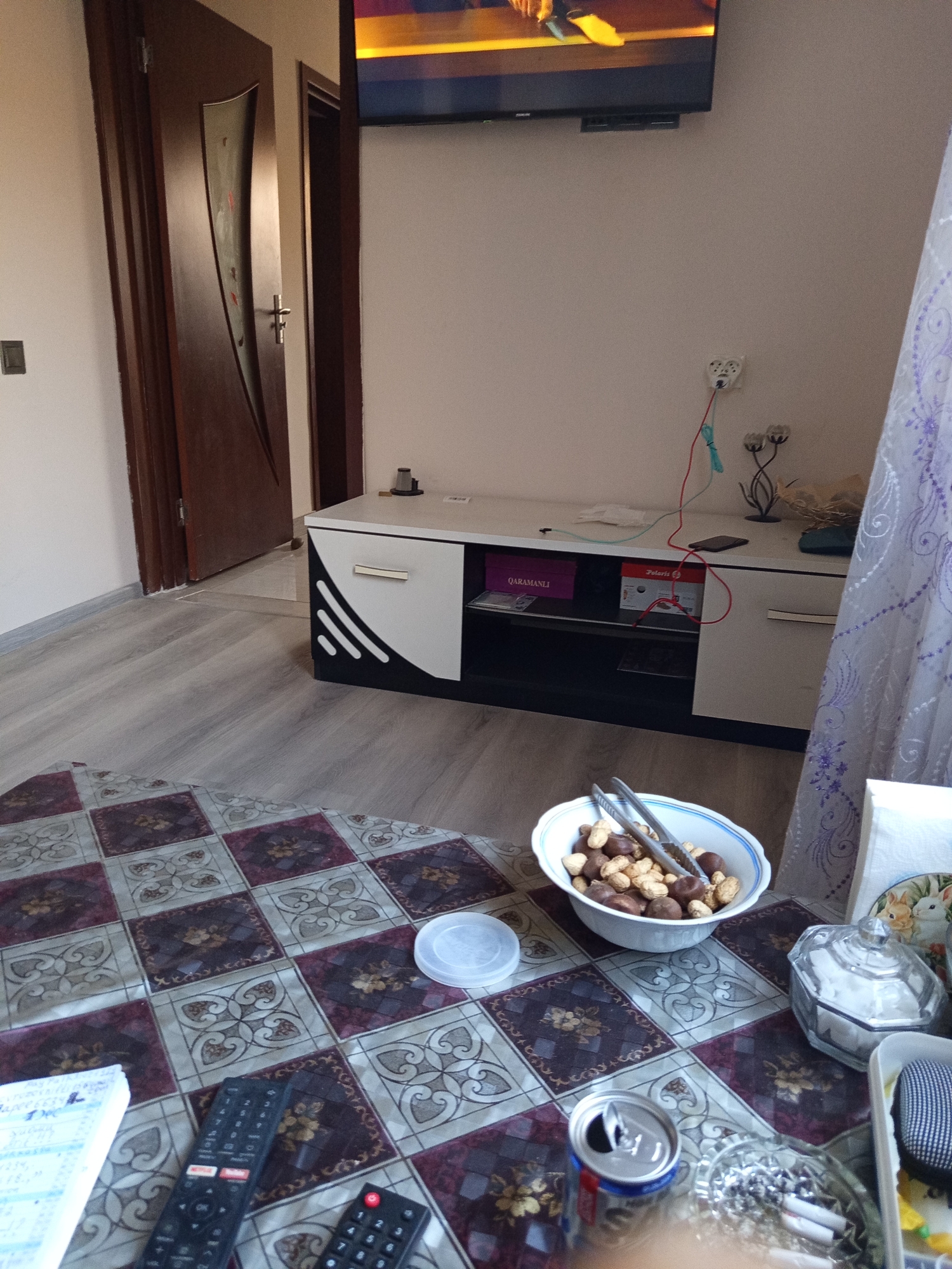 A living room area is depicted with a closed wooden door on the left, a TV mounted on the wall above a white and black entertainment unit, and a patterned rug covering part of the floor. On the entertainment unit, there are two boxes, a phone, and electrical cords. In the foreground, a bowl of nuts with tongs sits on the rug, next to an open soda can and an ashtray with cigarette butts. Other objects visible include a notebook, two remote controls, a lidded container, and a decorative glass jar filled with sugar cubes. The overall scene suggests a relaxed, indoor setting, possibly in a residential apartment in Shaghan, Azerbaijan.