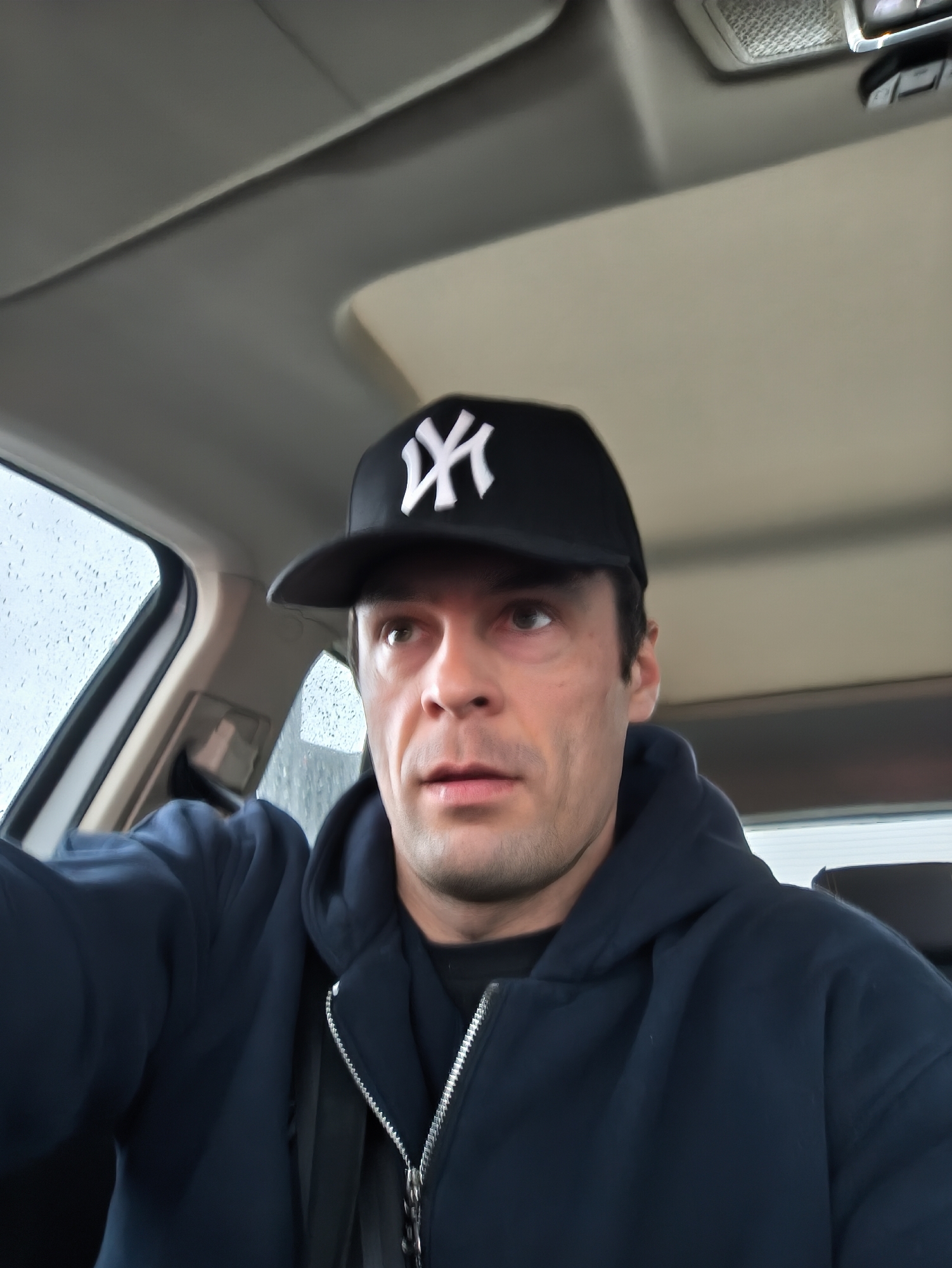 A man wearing a black baseball cap with a white "NY" logo and a blue zip-up hoodie is inside a car. Raindrops are visible on the car's window. The interior of the car is gray, and a portion of the sunroof is visible. The man appears to be taking a selfie. The location cannot be determined to be Moscow, Russia from this image alone.