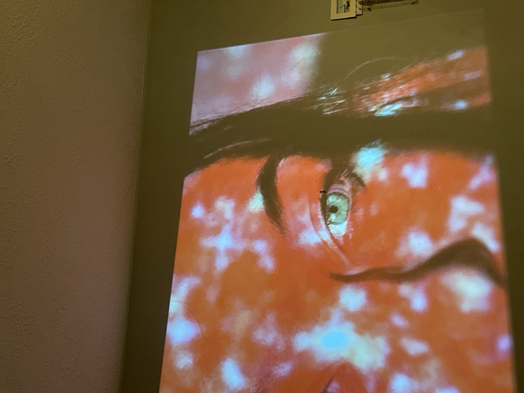 The image shows a close-up of a face with one visible eye, projected onto a wall. The face is predominantly orange and red, with blurred light spots scattered across it. The eyebrow above the visible eye is dark and prominent. The eye itself is greenish. The wall is a light, matte color, and there are some small framed objects near the top right corner. The location in Maidstone, United Kingdom is not apparent from this image.
