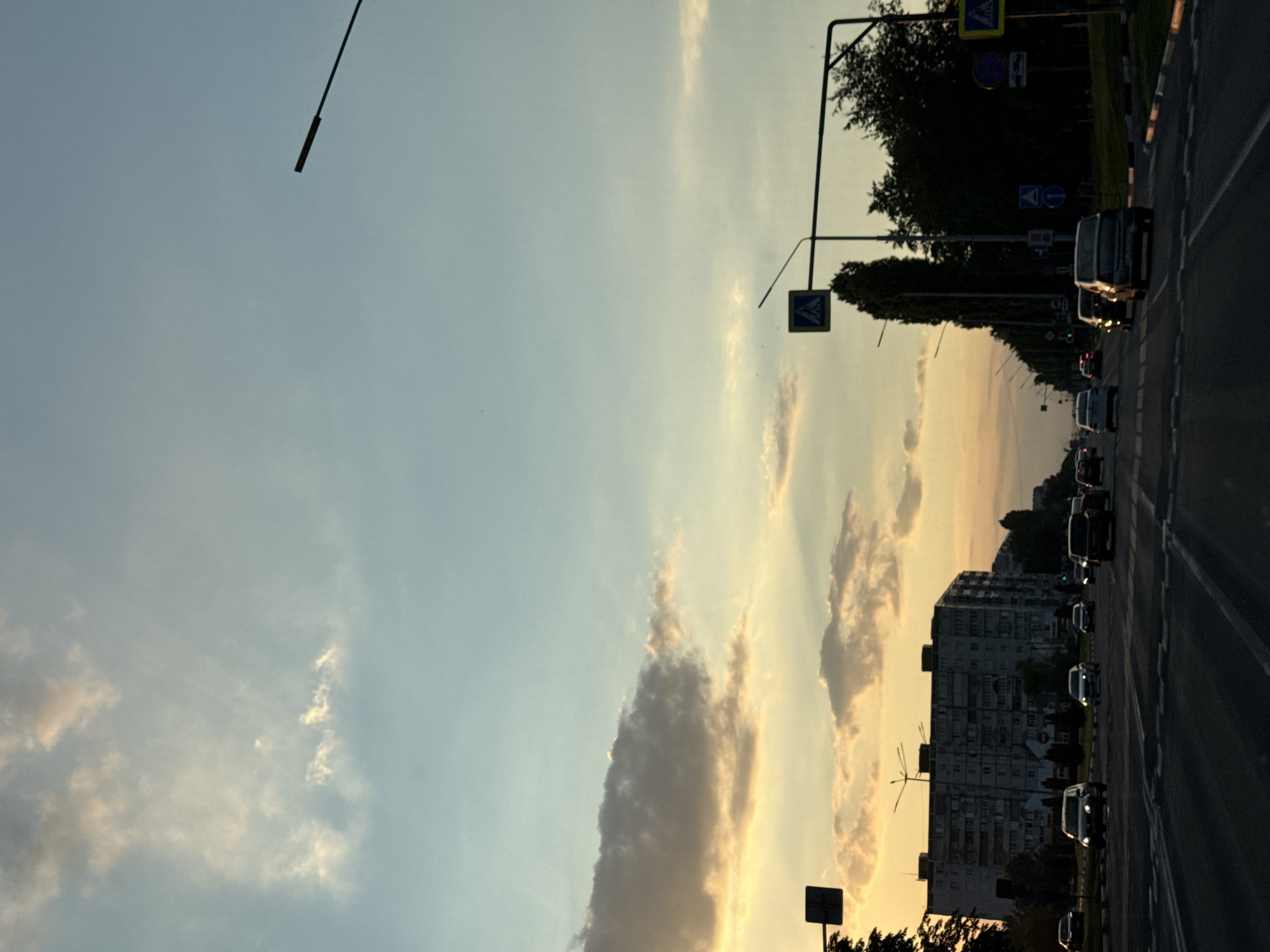 The image depicts a street scene at dusk in Stary Oskol, Russia. The sky is a blend of pale blue and sunset orange, with scattered clouds. A multi-story residential building, typical of Soviet-era architecture, dominates the left side of the frame, silhouetted against the fading light. Cars are visible on a multi-lane road in the foreground, with some headlights illuminated. Traffic lights and pedestrian crossing signs are present along the street. Trees line the right side of the road, forming a dark backdrop. The overall impression is of a functional urban environment transitioning into evening.