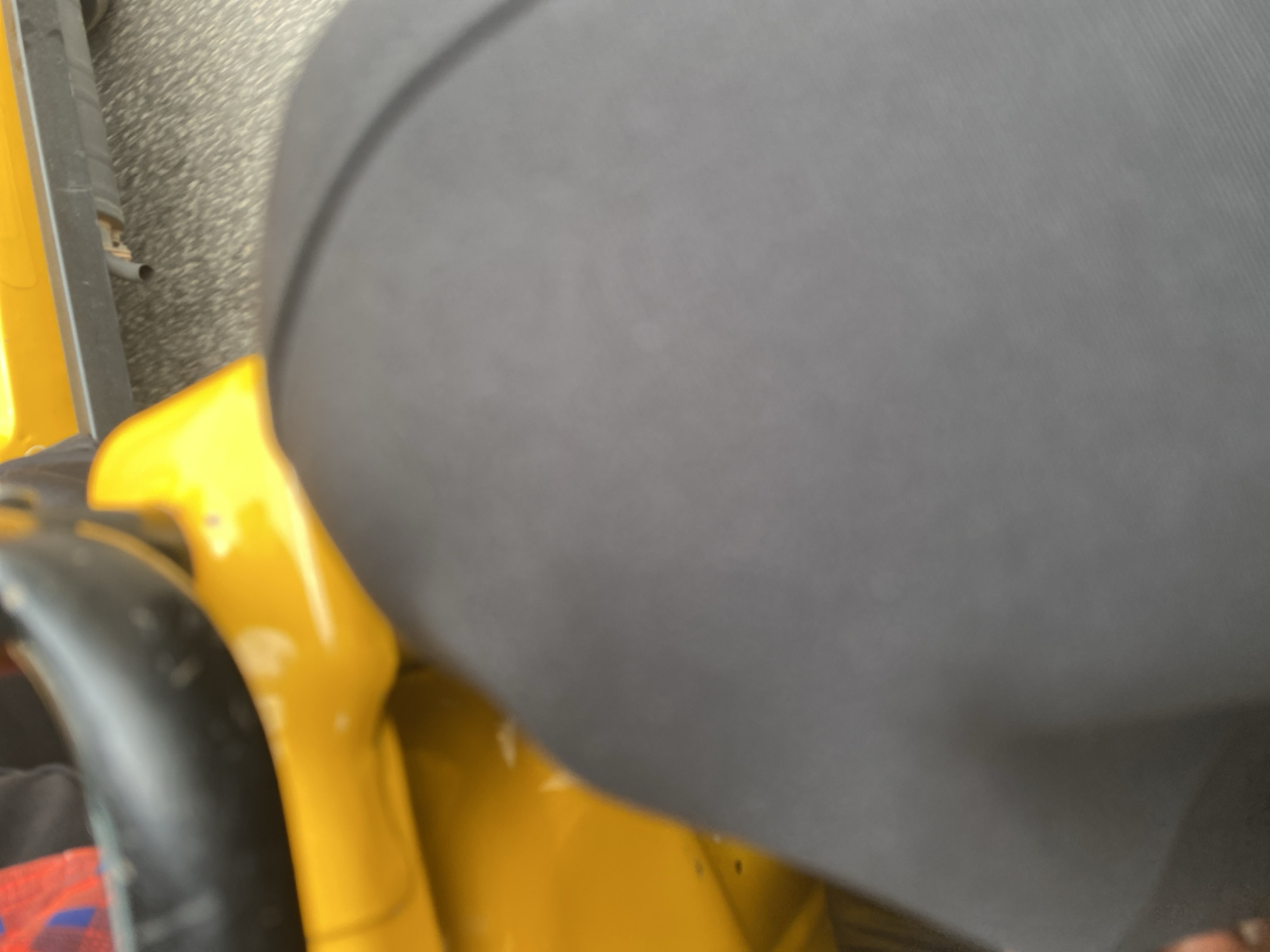 The image shows a close-up view of a person sitting on a yellow motorized tricycle, commonly known as an "all-in-one" or Keke Napep. The person is wearing dark-colored clothing, possibly leggings, which cover a significant portion of the frame. The yellow body of the tricycle is visible, along with a black handle or frame element in the foreground. The background appears to be a textured surface, possibly asphalt or concrete, suggesting an outdoor setting. The lighting suggests it might be daytime, but the specific time of day or weather conditions are not discernible. There are no visible text elements in the image. The focus is on the texture of the clothing and the bright yellow of the tricycle, with a shallow depth of field blurring the background. The scene is likely taking place on a street or road in Jalingo, Nigeria, given the context provided.