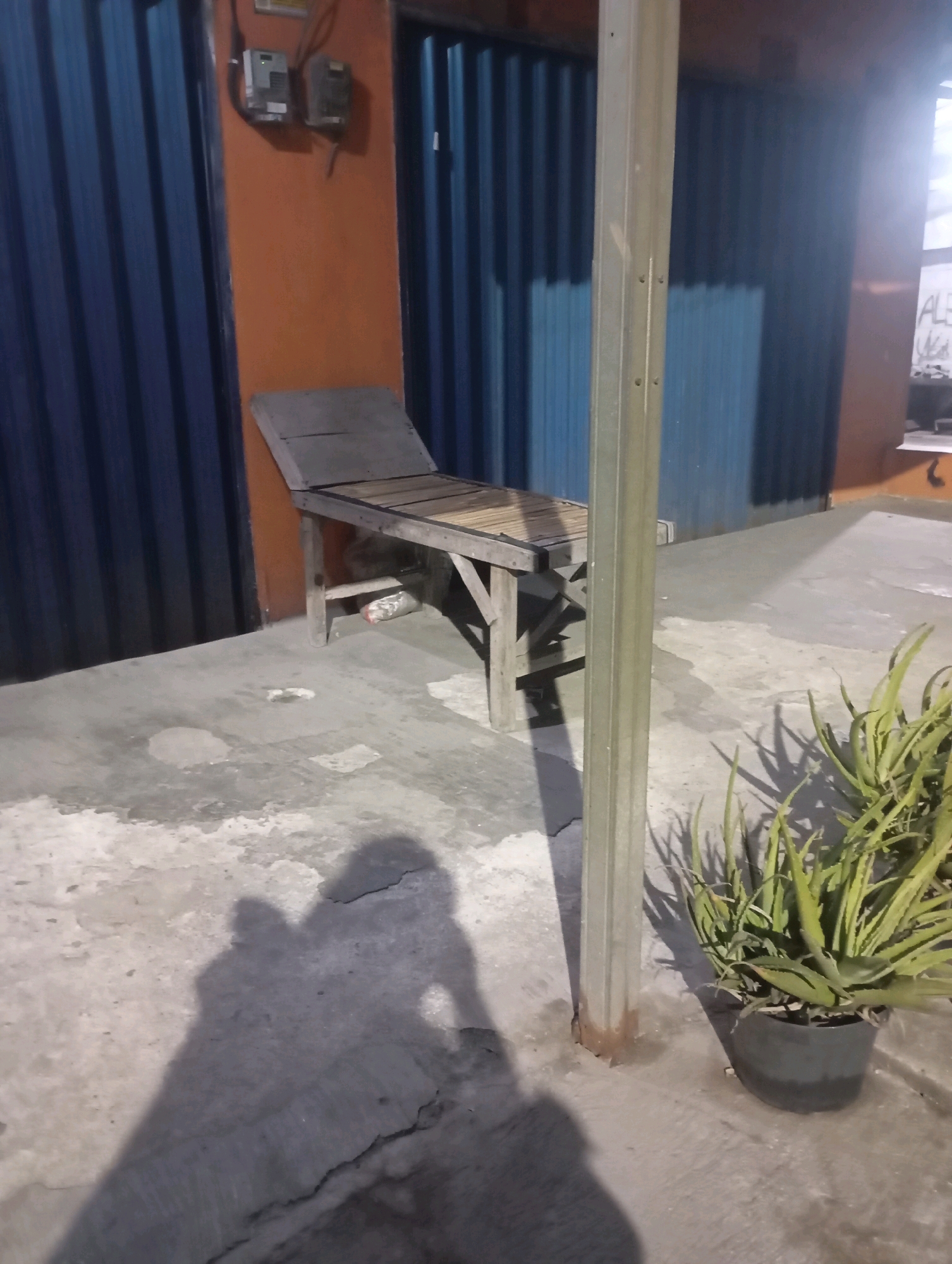In Babelan, Indonesia, a wooden bench sits on a concrete patio in front of a building with blue metal siding and orange-painted walls. Electrical boxes are mounted on the orange wall. A metal post stands vertically on the patio, and a potted plant with spiky green leaves is located near the post. The ground is wet in places, and shadows of two people are visible on the concrete.