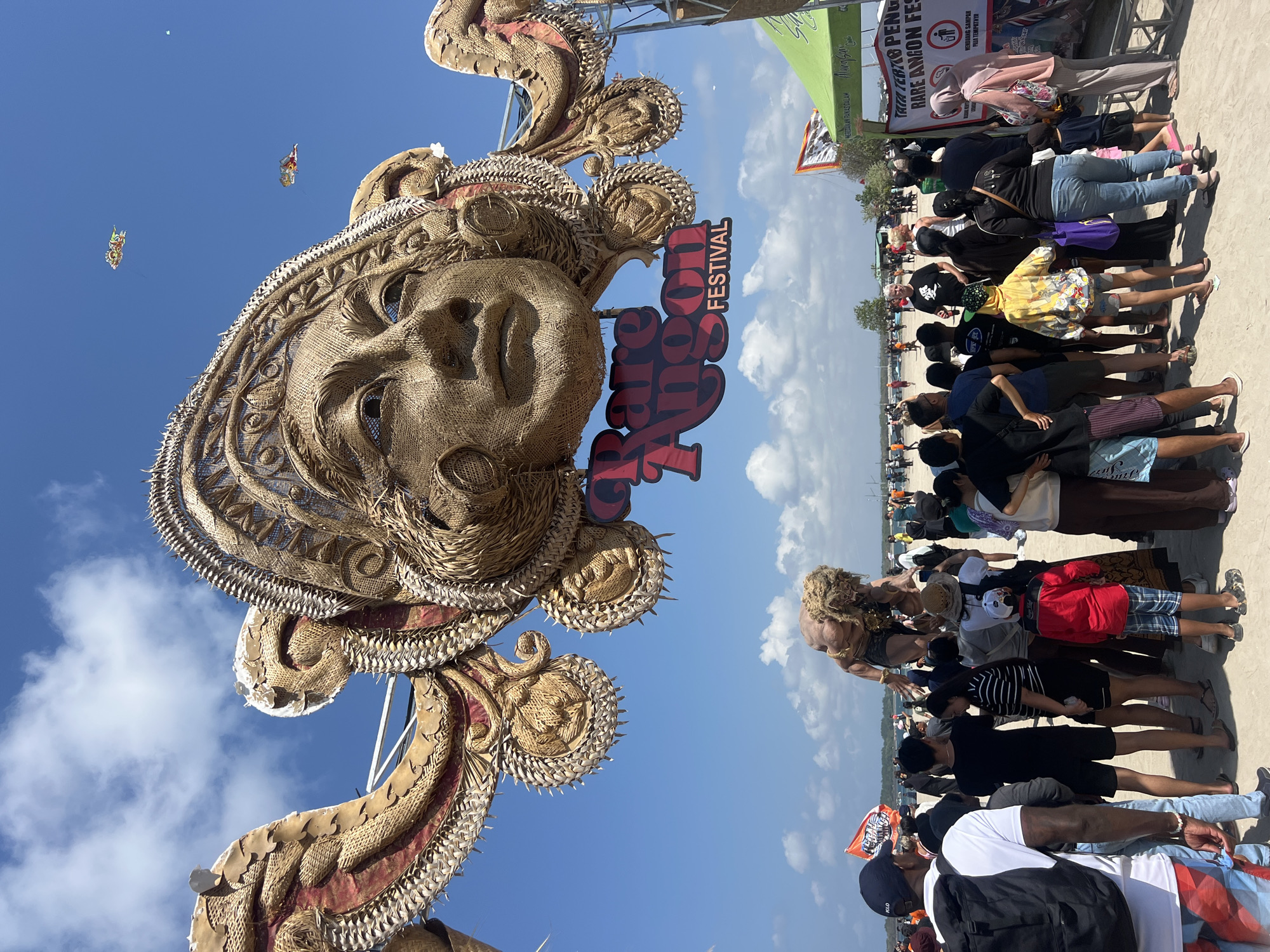 An outdoor festival is being held on a beach, likely in Denpasar, Indonesia. A massive, intricately crafted human face sculpture made of natural materials like straw and wood is the centerpiece, adorned with ornate ear-like structures and a headdress. Below the face, a sign reads "Rare Angon Festival." In the background, a large, muscular figure with long blonde hair appears to be posing or performing. A crowd of people of various ages and ethnicities are gathered, some observing the sculptures and performers, while others are milling about. Some people are wearing casual beach attire. There are also tents and banners, suggesting commercial or organizational presence at the event. In the sky, kites are visible, adding to the festive atmosphere. The environment is sunny with scattered clouds.