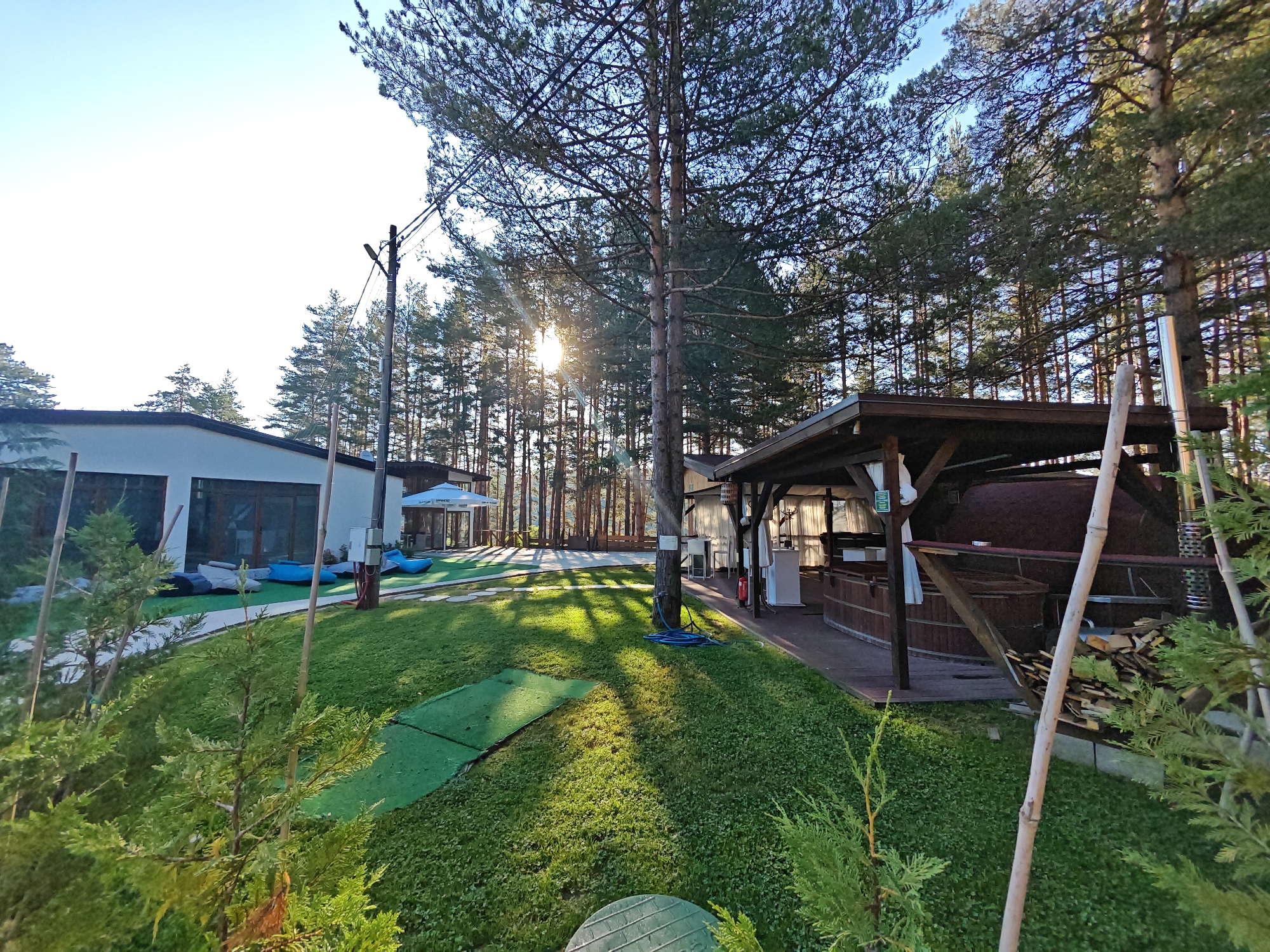 This media file depicts an outdoor scene at a resort or similar establishment, likely in Alino, Bulgaria. The setting is a well-maintained grassy area with several pine trees and some younger evergreen shrubs. In the background, a modern, white building with large windows is visible, along with a white umbrella structure and some blue inflatable seating. To the right, there's a wooden structure with a covered patio area, featuring what appears to be a wood-fired oven and a stack of firewood.
The time of day appears to be late afternoon or early morning, with the sun casting long shadows across the lawn and creating a warm, dappled light effect through the trees. The sky is clear and blue, suggesting pleasant weather.
There is no visible activity or event taking place. The scene is static, conveying a sense of tranquility and a potential for outdoor leisure. The location cues, such as the architecture and the surrounding pine forest, suggest a natural, possibly mountainous or hilly, environment. The overall mood is peaceful and inviting. No discernible text is visible in the image.