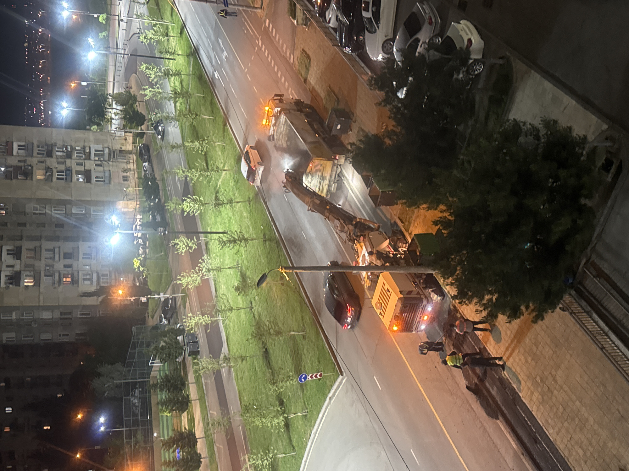 The image shows an urban scene at night. A road stretches diagonally across the frame, separating a grassy median strip with young trees on one side, and a sidewalk with trees adjacent to a building on the other. Streetlights illuminate the scene.
On the right side of the image, a crane and a utility truck with flashing lights are parked on the road. Two individuals wearing reflective vests are standing on the sidewalk near the truck. A black car is parked in front of the utility truck, facing away from the camera.
On the other side of the road, a garbage truck with flashing lights and a white car behind it are visible.
The location is likely Tbilisi, Georgia, based on architectural styles and street design. The scene appears to be a public works operation underway at night.
