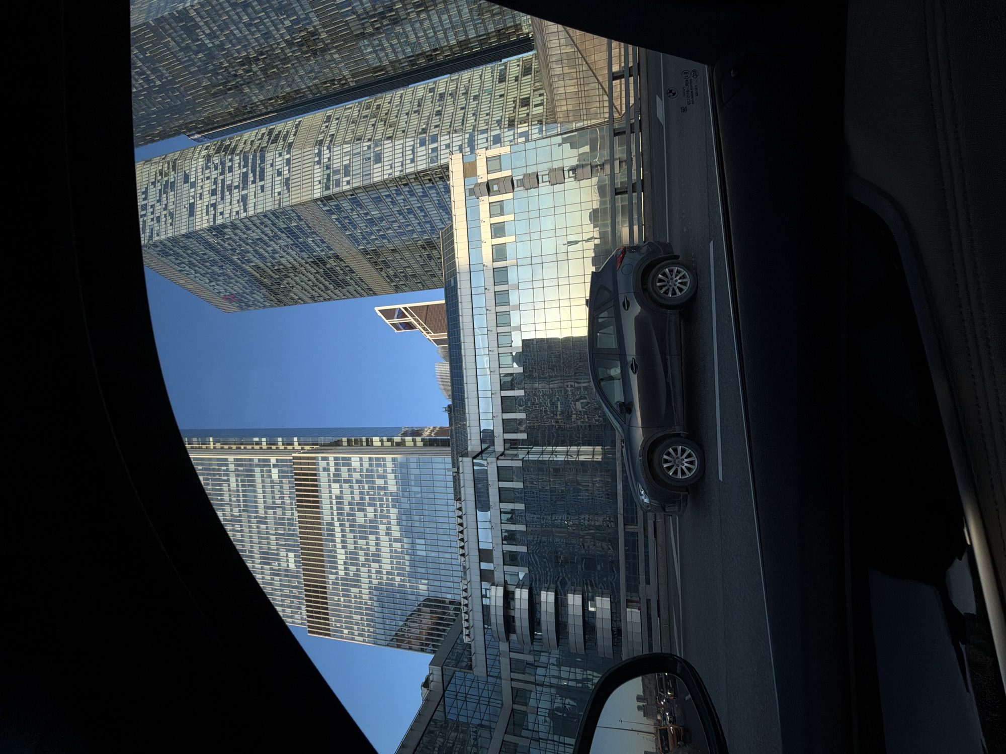 The image, taken from inside a vehicle, shows the Moscow International Business Center, also known as Moscow City. Skyscrapers fill much of the frame, reflecting light and appearing as tall glass-clad structures. A grey car is visible on the road adjacent to the buildings. The sky is clear and blue. The interior of the vehicle is dark, with parts of the car's interior visible along the edges of the frame, including the side mirror.