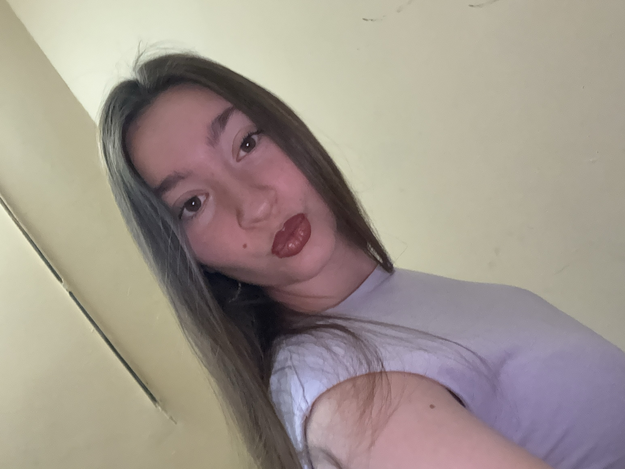 The image is a low-angle, close-up selfie of a young woman with long, brown hair and dark eyes. She is wearing a light purple t-shirt. She has on lipstick and a mole above her upper lip on the right side of her face. The background is a plain, light-colored wall and ceiling, suggesting an indoor setting. The lighting is warm and appears to be artificial. There are no discernible landmarks or features that would indicate a specific location within the city of Sofia, Bulgaria.