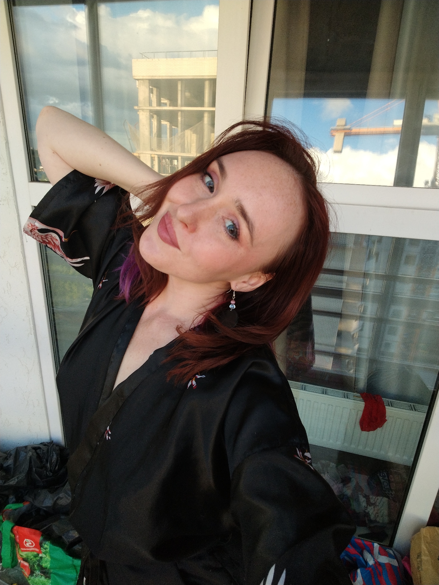 A selfie captures a woman with reddish-brown hair, fair skin, freckles, and blue eyes, smiling softly at the camera. She wears a black, satin-like robe or kimono patterned with white and red birds, and drop earrings featuring blue and pink beads. Her left arm is raised with her hand behind her head.
The setting is indoors, likely a balcony or a room with a large window, overlooking an urban environment in Izhevsk, Russia. The window reflects a cloudy sky with patches of blue. Through the glass, a multi-story concrete building under construction and an orange construction crane are visible against the sky.
In the foreground, to the woman's right (viewer's left), several black plastic bags and a green bag with a red, indistinct logo are piled. Below the window, a white radiator and a piece of red fabric are seen. The lighting suggests daytime. The woman appears relaxed and pleasant. No discernible text is visible on the green bag.