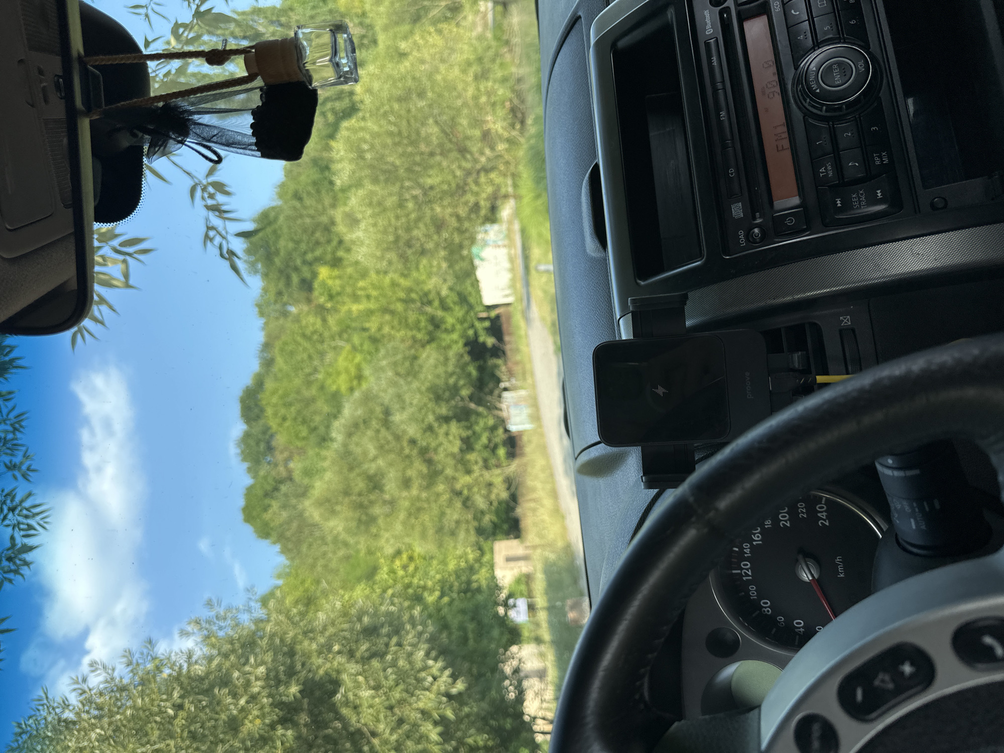 The image shows the interior of a car, looking out through the windshield at a rural landscape. The dashboard is visible, with the steering wheel on the left and the car's radio on the right. A phone mount with a charging phone is attached to the dashboard. Outside the car, there is a road, trees, and a cloudy blue sky. The overall impression is a daytime scene. No people are visible in the image. The location is the village of Makiv, Ukraine, as indicated by the background details of rural buildings and vegetation consistent with the area.