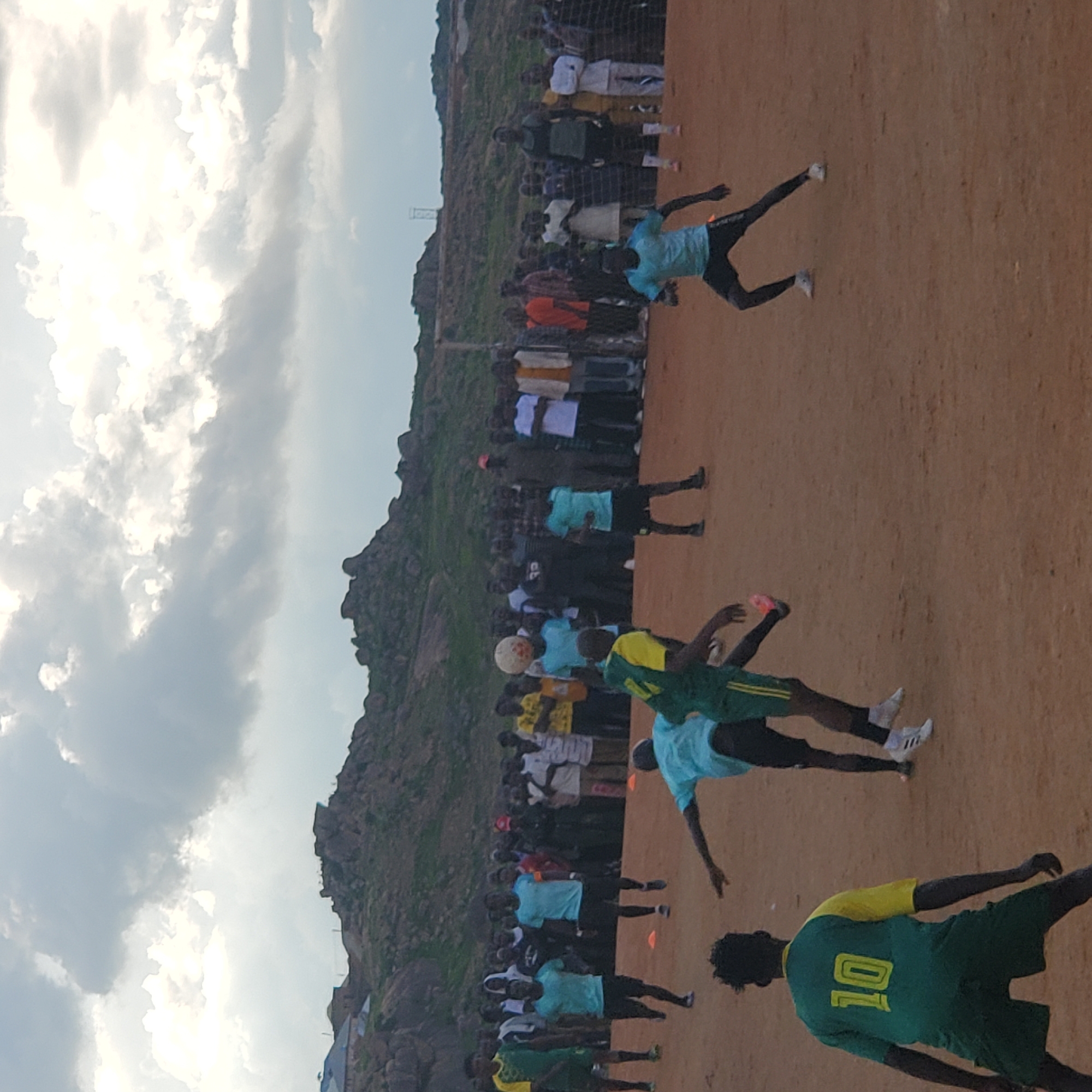 The image depicts a scene in Jos, Nigeria, featuring an outdoor soccer match on a dirt field. A crowd of spectators stands behind a net, observing the game. Several players are actively engaged on the field, with one player in the foreground wearing a green and yellow jersey with the number "10". Another player in a light blue jersey is in motion, seemingly about to kick a soccer ball. The background features rocky hills and a cloudy sky, characteristic of the topography around Jos. The overall atmosphere suggests a local sporting event with community participation.