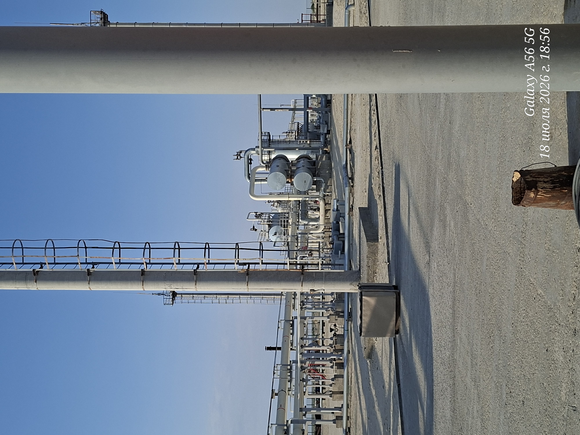 This media file depicts an industrial scene featuring large-diameter pipes and complex piping systems, likely part of a refinery or chemical plant. The scene is viewed from a low angle, emphasizing the scale of the industrial infrastructure against a clear blue sky. Multiple large, grey, cylindrical pipes run horizontally across the frame, supported by structures. One prominent pipe is positioned close to the viewer in the foreground. Below this, a more intricate network of smaller pipes, valves, and industrial equipment is visible. There are metal walkways and safety railings associated with the piping systems, suggesting an operational facility.
The lighting indicates it is daytime, with shadows cast by the structures, suggesting the sun is at a moderate angle. The overall impression is one of a functional, industrial environment.
Visible text on the right side of the image reads: "Galaxy A56 5G" and "18 июля 2026 г. 18:56". This text, likely a timestamp and device identifier, is superimposed on the image.