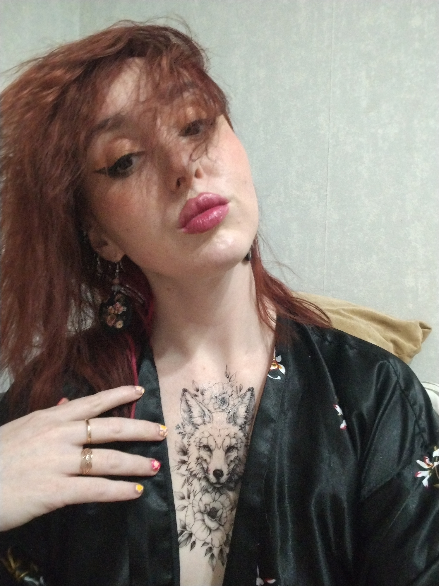 A young woman with red hair and fair skin poses for a selfie. She wears a black kimono with floral patterns. A tattoo of a fox and flowers is visible on her chest. She has on makeup, including winged eyeliner and glossy pink lipstick. She wears dangling earrings and has two rings on her left hand. Her fingernails are painted with different designs. The background appears to be a plain, light-colored wall. It is not possible to verify the location in the city of Izhevsk, Russia from the image.