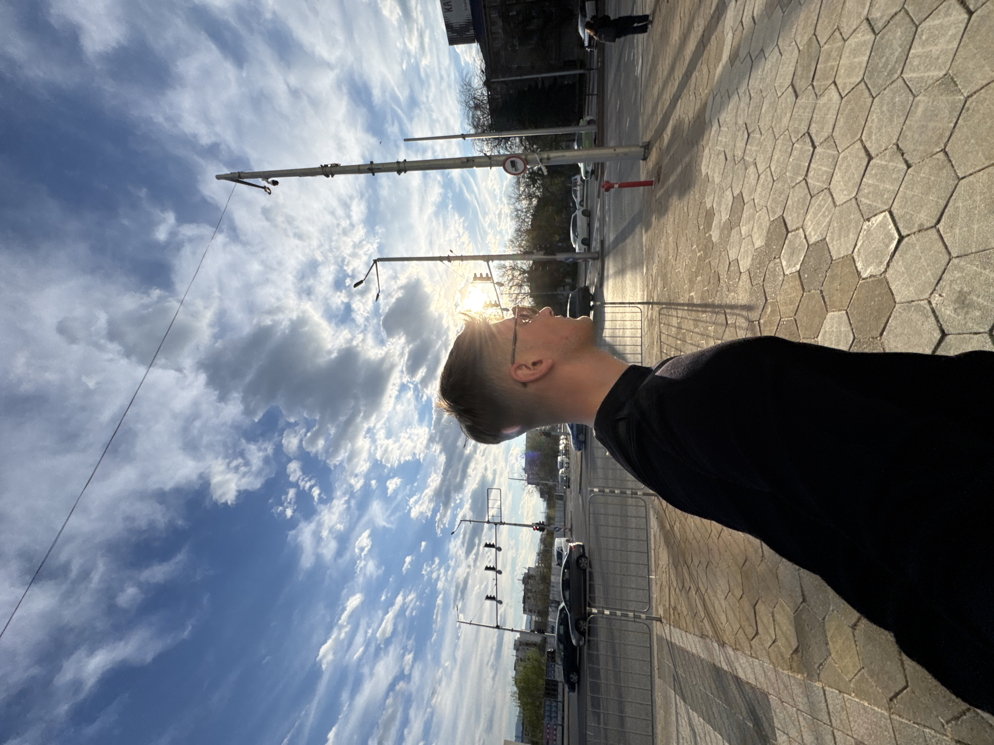 A photograph of a man in Plovdiv, Bulgaria, taken from a low angle, captures the subject in the right foreground with his head turned upward, facing toward the sun and sky. The sky is partially covered with white clouds against a blue backdrop. A street scene unfolds beyond the man, showing city infrastructure including traffic lights, street lamps, and road signs. Cars are parked and moving along the street. The ground is paved with a hexagonal tile pattern.